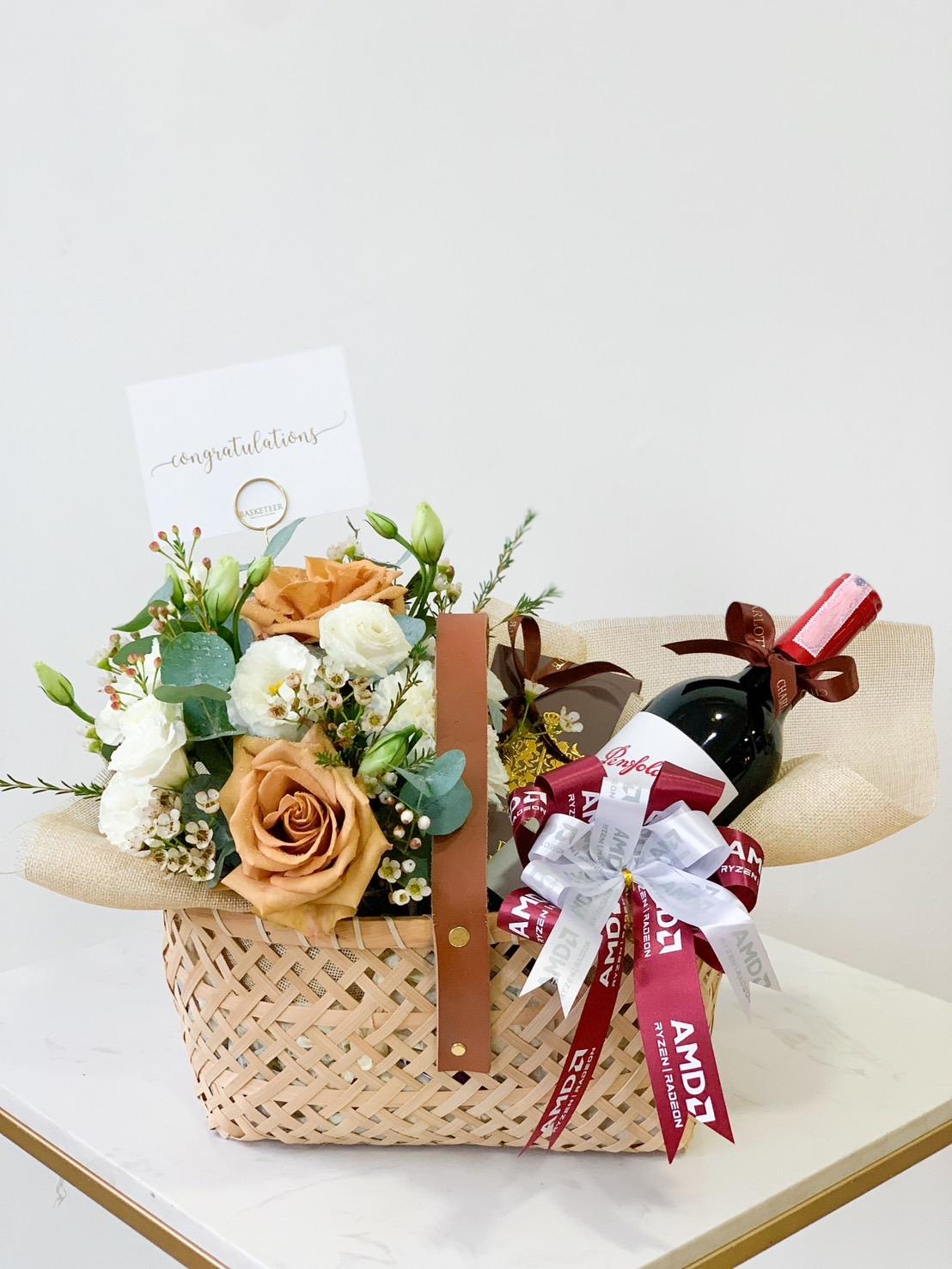 A wicker gift basket featuring a bouquet of white and peach roses, greenery, a bottle of wine adorned with a red bow, and a "Congratulations" card. The basket is placed on a white surface against a plain background.