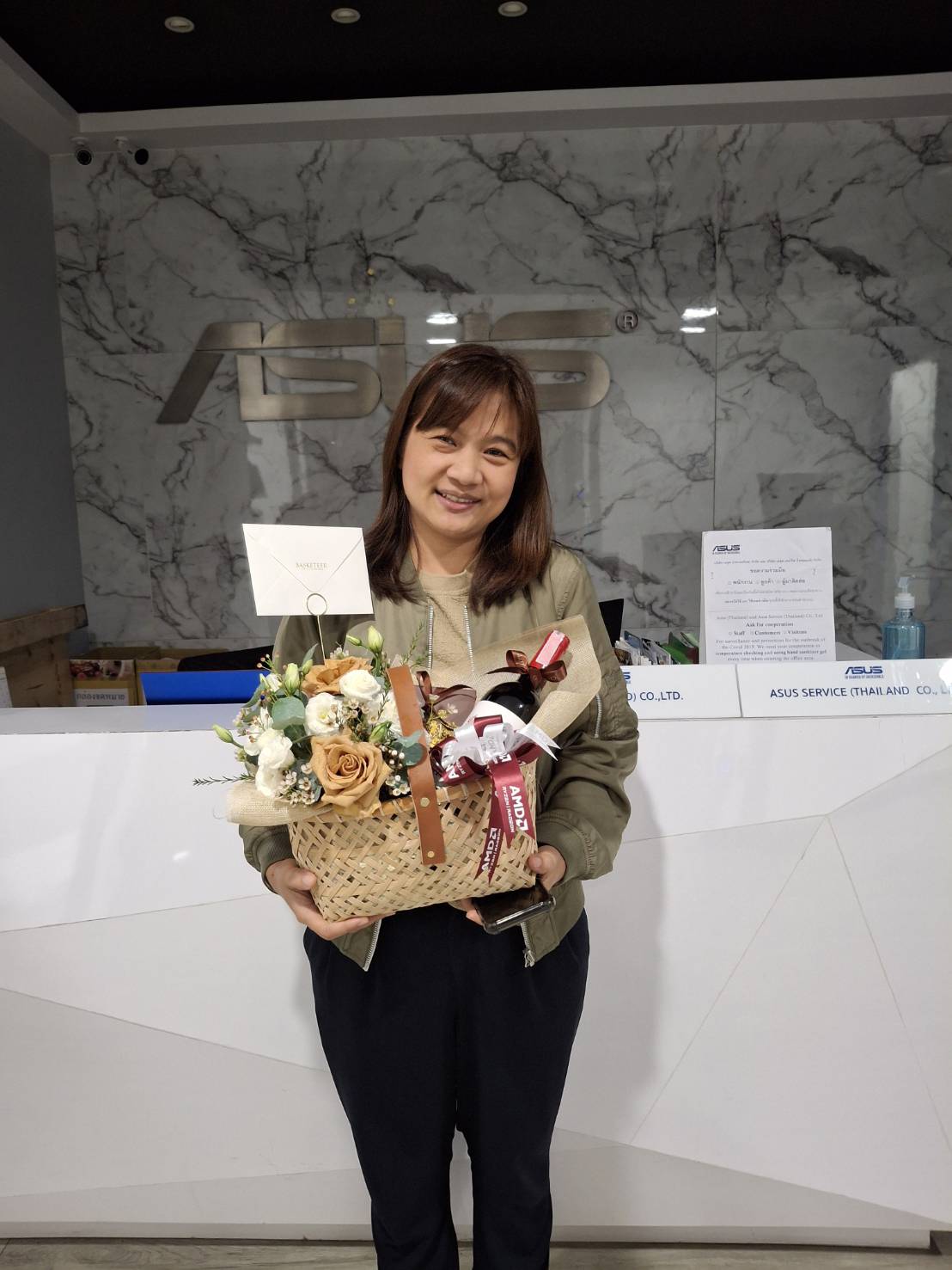 A woman with shoulder-length hair stands smiling inside an office with a marble wall in the background. She holds a gift basket filled with flowers and various items, with a card attached to it. The company logo is visible on the wall behind her.