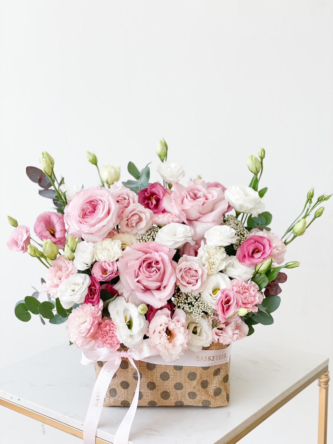 A large, elegant bouquet of pastel pink and white flowers, including roses and lisianthus, arranged in a polka-dotted beige basket. The flowers are lush and fresh, placed on a white surface against a plain light background.