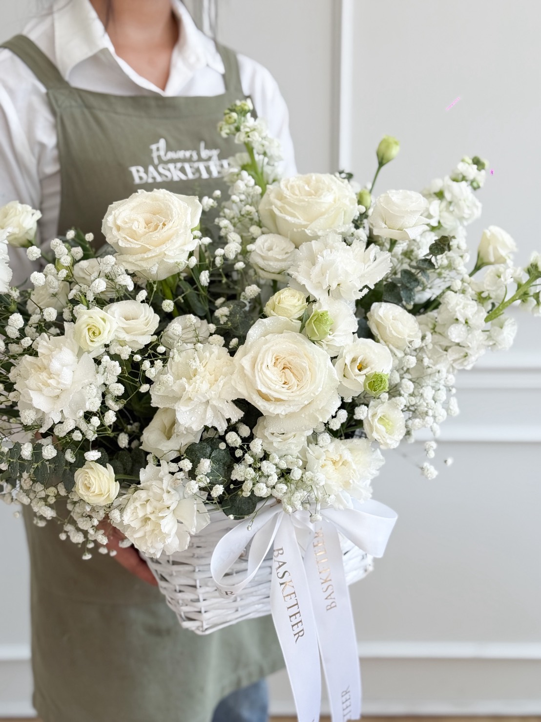 A person wearing a green apron holds a White Garden Serenity Flowers Basket filled with white roses, carnations, and baby's breath, adorned with a white ribbon. The softly lit, minimalistic background adds to the serene display.