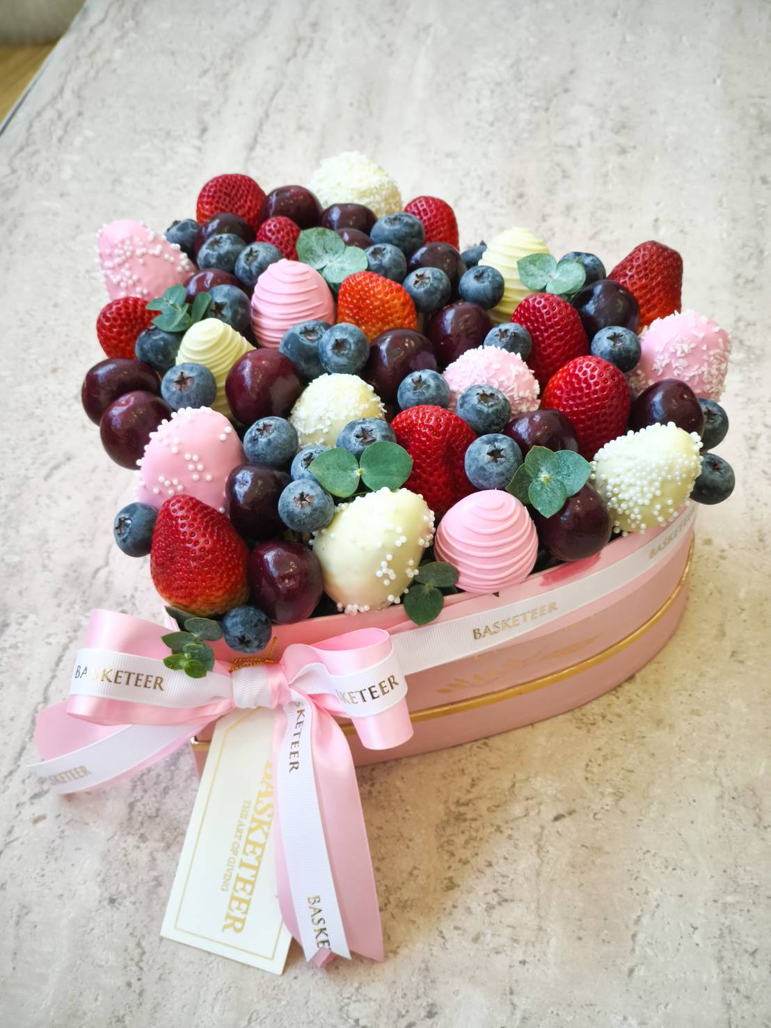 Heart-shaped gift box with chocolate-covered strawberries, blueberries, grapes, and fresh fruit, decorated with pink ribbons.