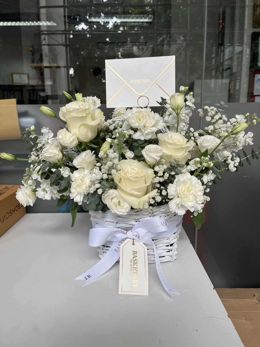A White Garden Serenity Flowers Basket filled with white roses, carnations, lisianthus, and baby's breath sits on a table. Adorned with a white ribbon and “BASKETTER” tag, an envelope is clipped above the flowers.