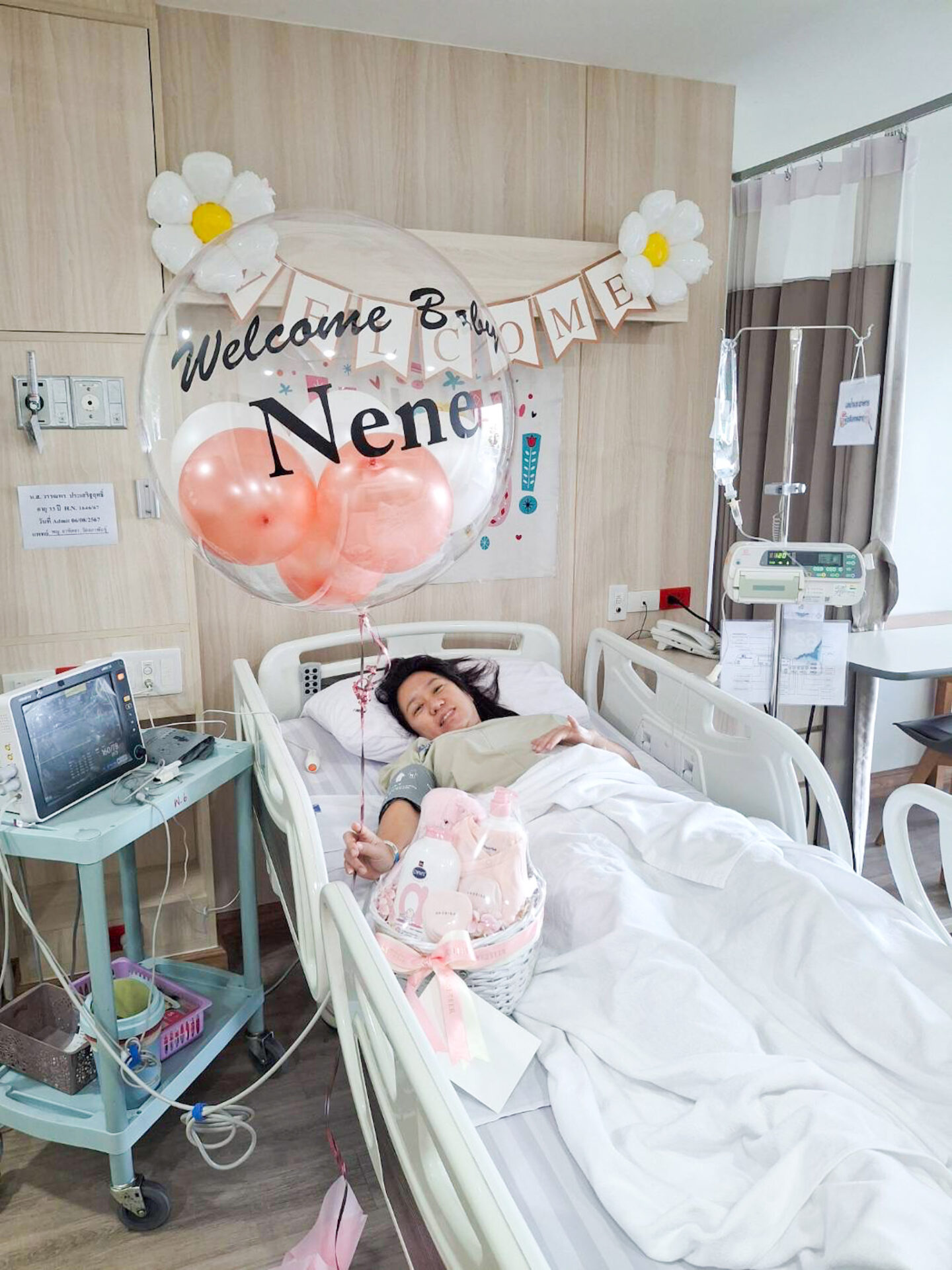 A woman lies in a hospital bed giving a thumbs-up, with a 