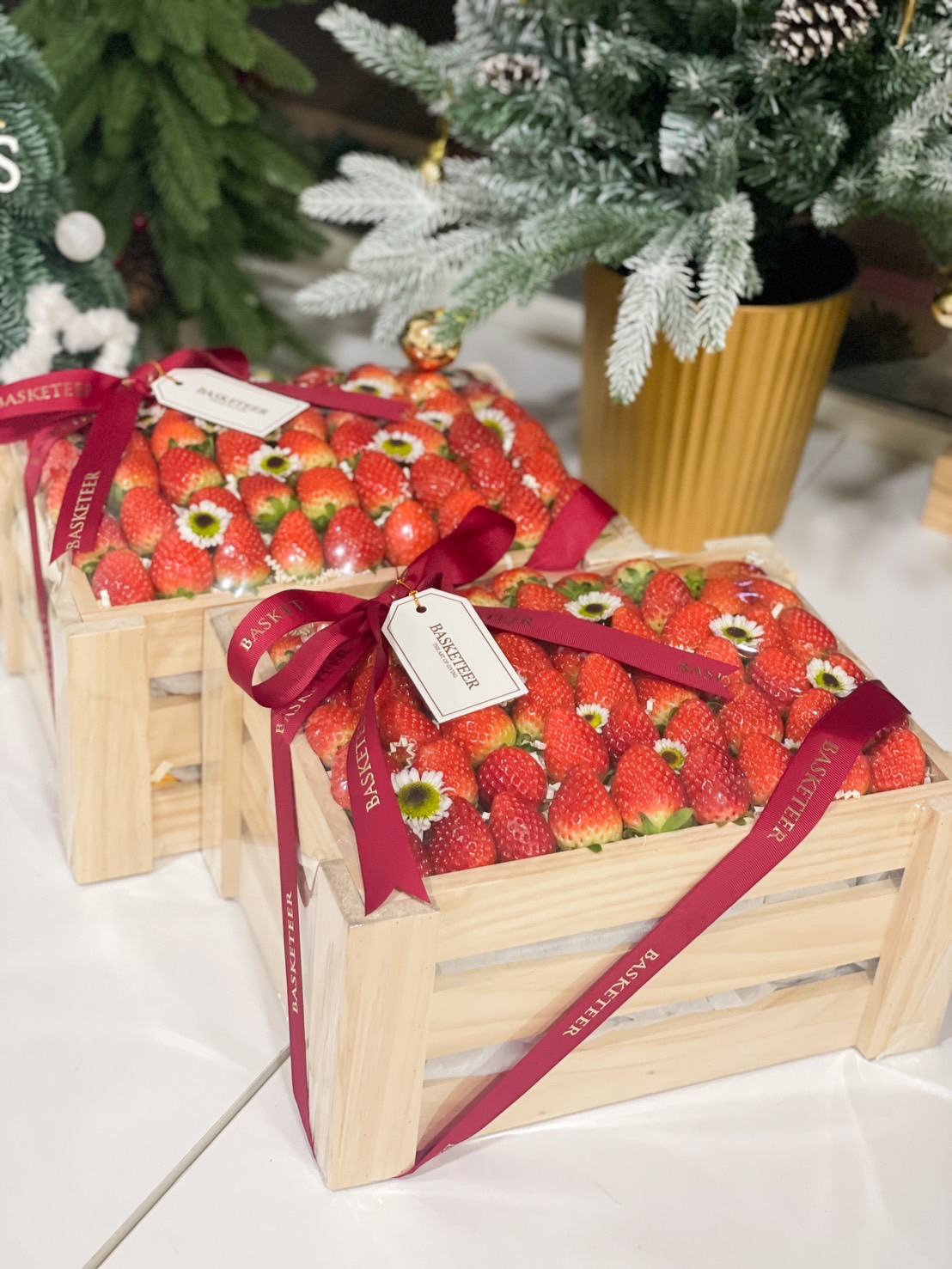 Rustic wooden crate filled with fresh, vibrant strawberries, adorned with white daisies and tied with a maroon Basketeer ribbon.