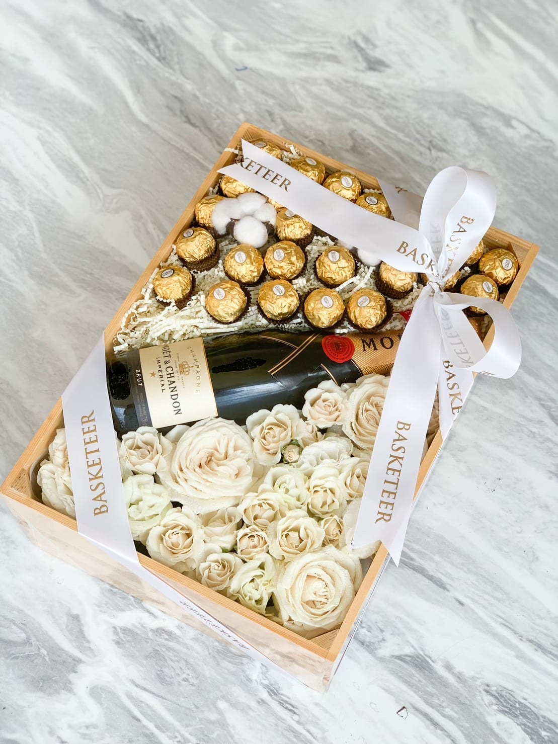 A Champagne & Chocolate With Flowers Gift Box, this wooden box contains a bottle of Moët & Chandon champagne, surrounded by white roses and Ferrero Rocher chocolates. Adorned with white ribbons labeled "BASKETEER" on a grey marbled surface, it exudes elegance.