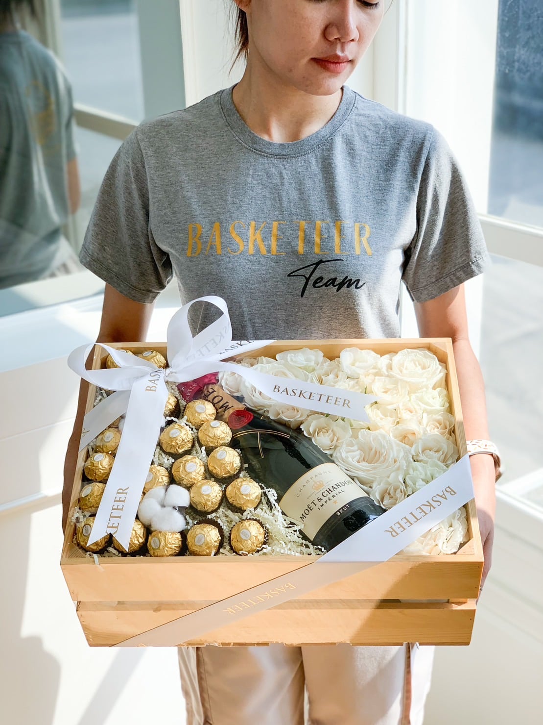 A person wearing a gray "BASKETEER Team" t-shirt is holding a wooden box filled with white roses, Ferrero Rocher chocolates, a bottle of Moët & Chandon champagne, and a white ribbon with "BASKETEER" printed on it—truly a Champagne & Chocolate With Flowers Gift Box.