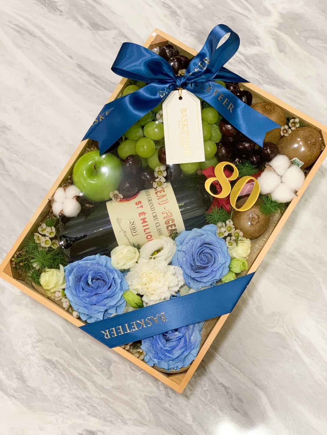 A luxurious wooden box elegantly brimming with succulent green grapes, ripe kiwis, plump cherries, and a crisp green apple, nestled among plush cotton flowers and an assortment of freshly bloomed roses. In the heart of this exquisite arrangement lies a sophisticated bottle of Merlot wine. Surrounding this enchanting scene are delicate berries and a charming ribbon adorned with the Basketeer logo, adding a touch of exclusivity. A nearby small glass dish filled with pearl-like ornaments enhances the opulent ambiance.