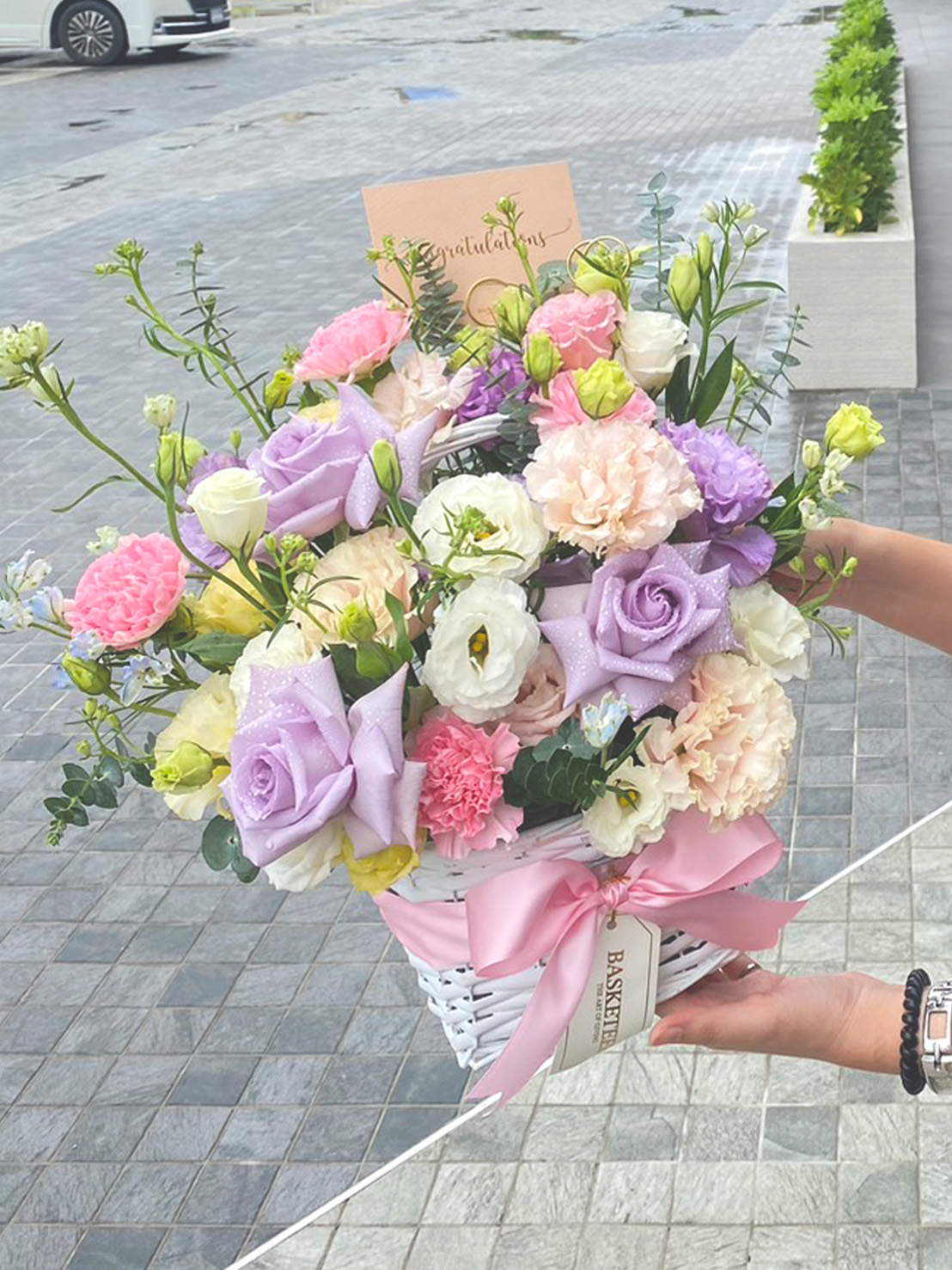 An elegant white wicker basket from Basketeer showcases a luxurious assortment of pastel blooms, featuring delicate pink, purple, and white roses alongside charming pink carnations and lush greenery. Adorned with an exquisite pink ribbon bow bearing the Basketeer logo, this arrangement rests on a pristine white marble surface against a sophisticated white paneled wall, creating an inviting and opulent display.