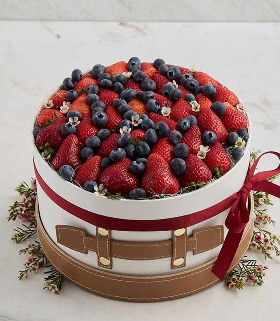 A round, exquisitely crafted cake, artfully designed to resemble a vintage suitcase, graces an elegant marble surface. This luxurious Berry Harvest Gift Box by Basketeer is adorned with a sumptuous red ribbon bearing the distinguished Basketeer logo. Brimming with an assortment of fresh, luscious strawberries and plump blueberries, each bite promises a taste of pure indulgence. Delicate small flowers embellish the top while verdant greenery and charming pink blossoms encircle its base, creating an inviting tableau that exudes sophistication and opulence.