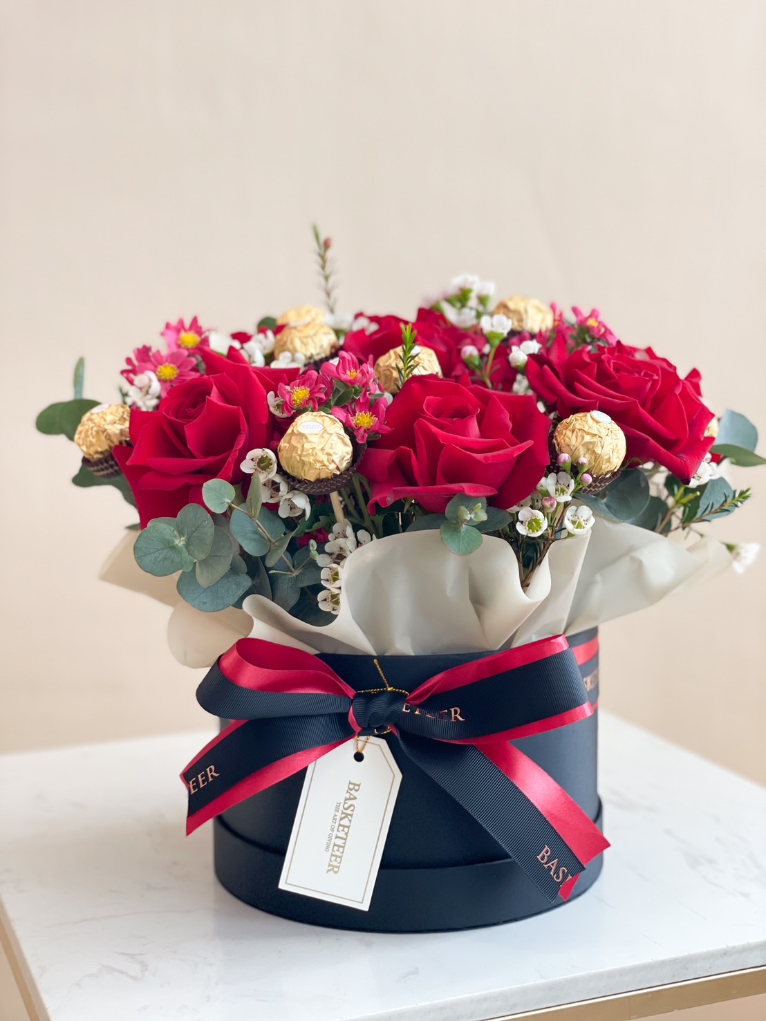 A luxurious floral gift box featuring red roses, Ferrero Rocher chocolates, and delicate floral accents, arranged in a navy round box with a satin ribbon.