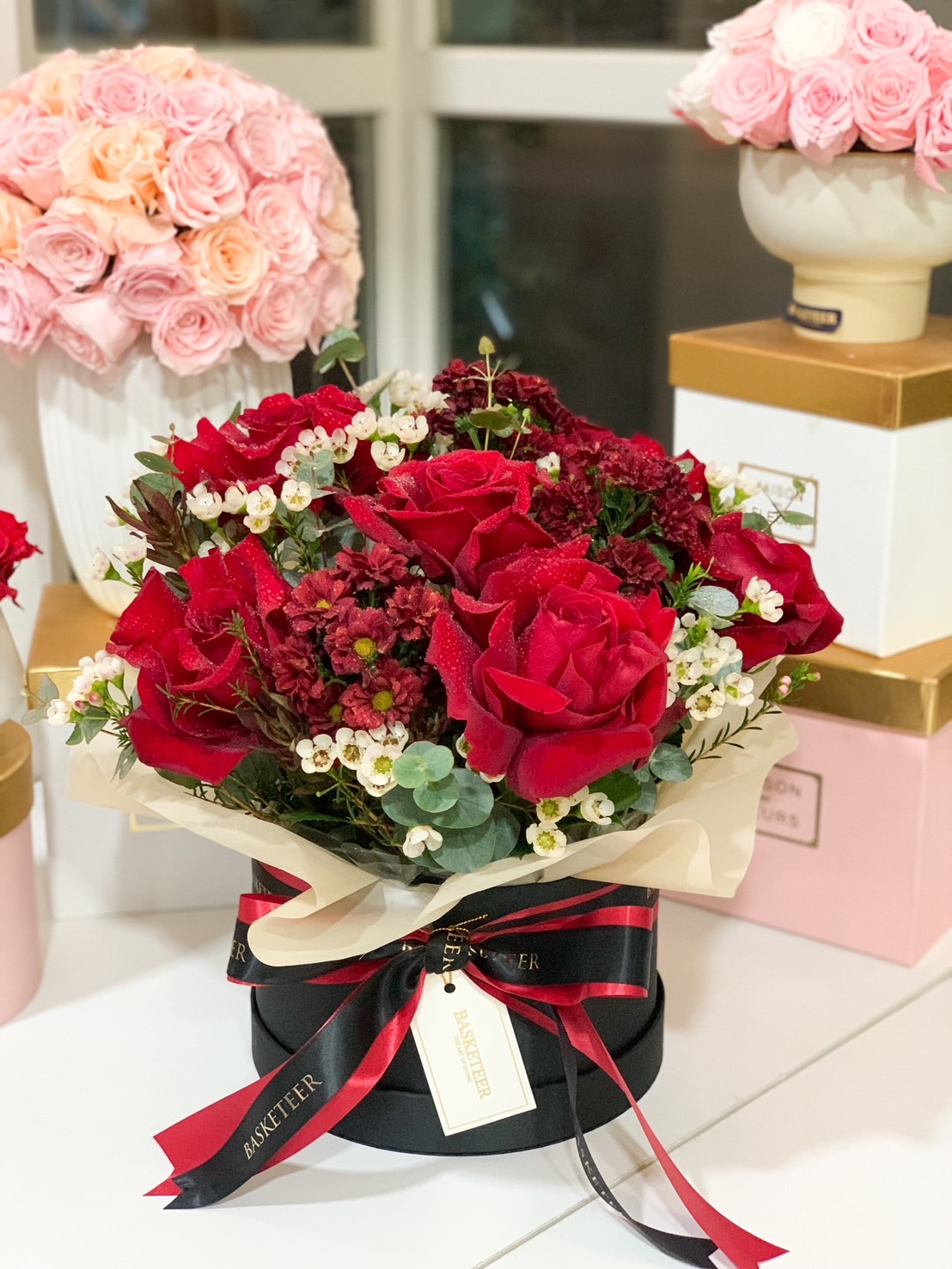 A luxurious black floral box filled with vibrant red roses, white wax flowers, and elegant greenery, tied with a black and red satin ribbon.