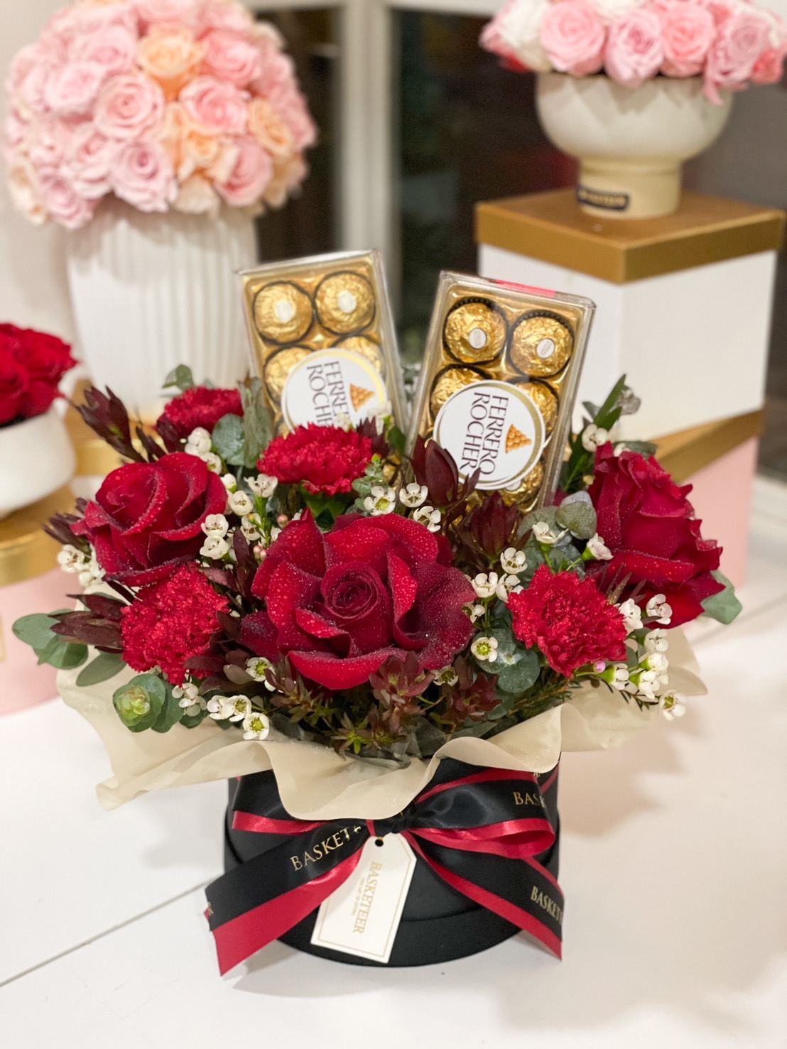 A luxurious gift box featuring fresh red roses, Ferrero Rocher chocolates, and delicate white flowers, tied with a black and red satin ribbon.