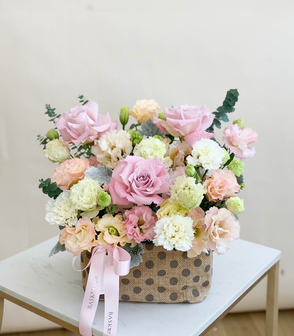 Elegant floral arrangement in a polka dot fabric basket featuring pink, peach, and white roses with eucalyptus leaves and a pink ribbon.