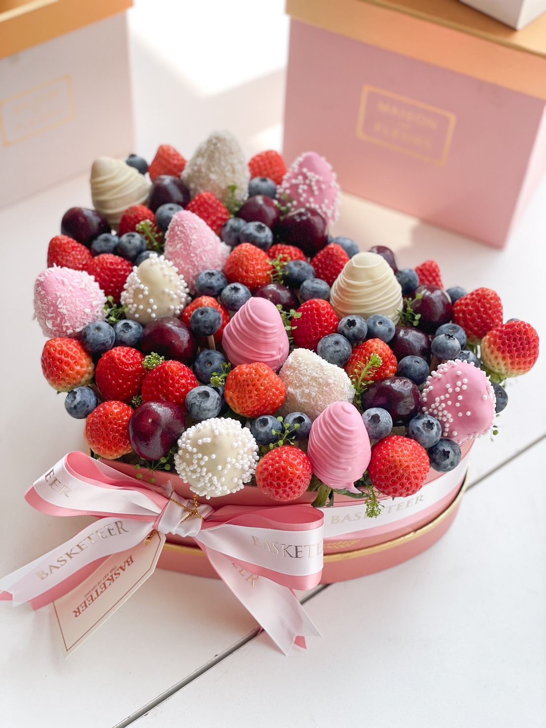 A heart-shaped box filled with fresh strawberries, blueberries, cherries, and elegant chocolate-covered berries, beautifully wrapped with a satin ribbon.