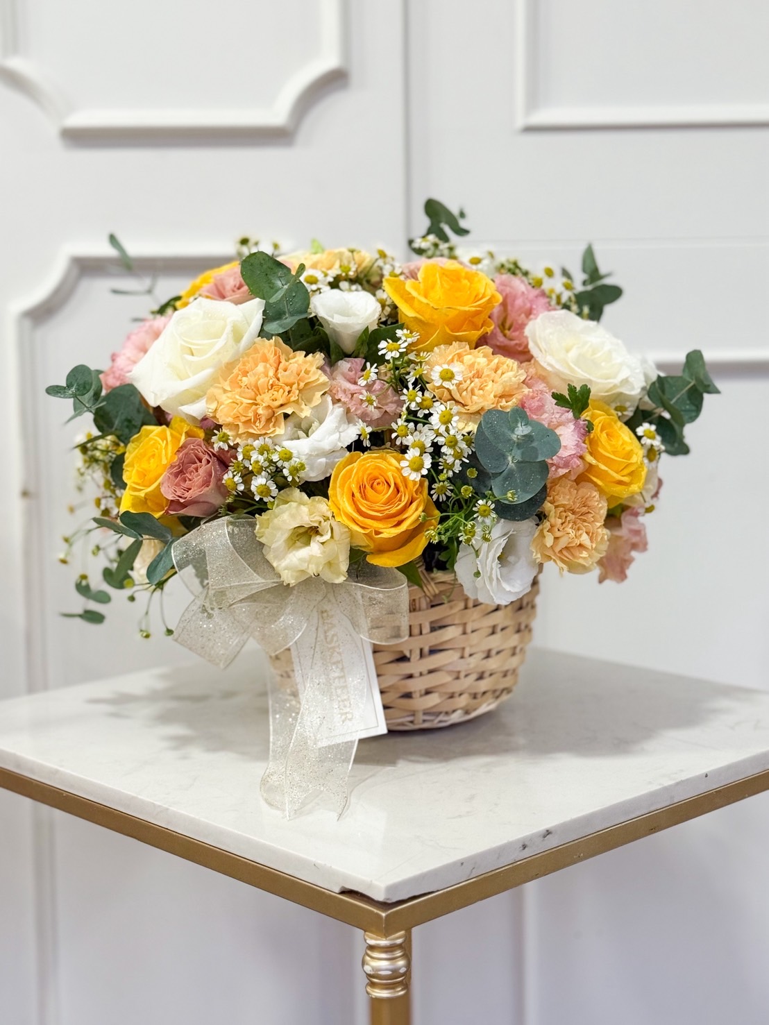 Bright flower basket with yellow roses, white roses, peach carnations, pink lisianthus, chamomile flowers, and eucalyptus.