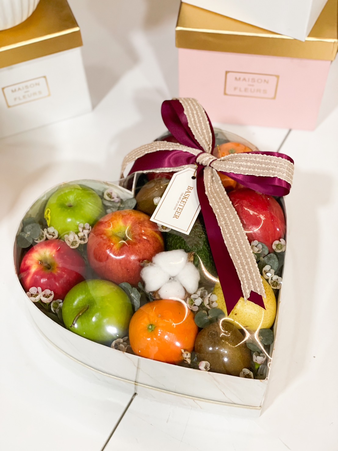 A heart-shaped transparent fruit box filled with fresh apples, oranges, kiwis, and pears, decorated with flowers and tied with a luxurious ribbon—perfect for gifting.
