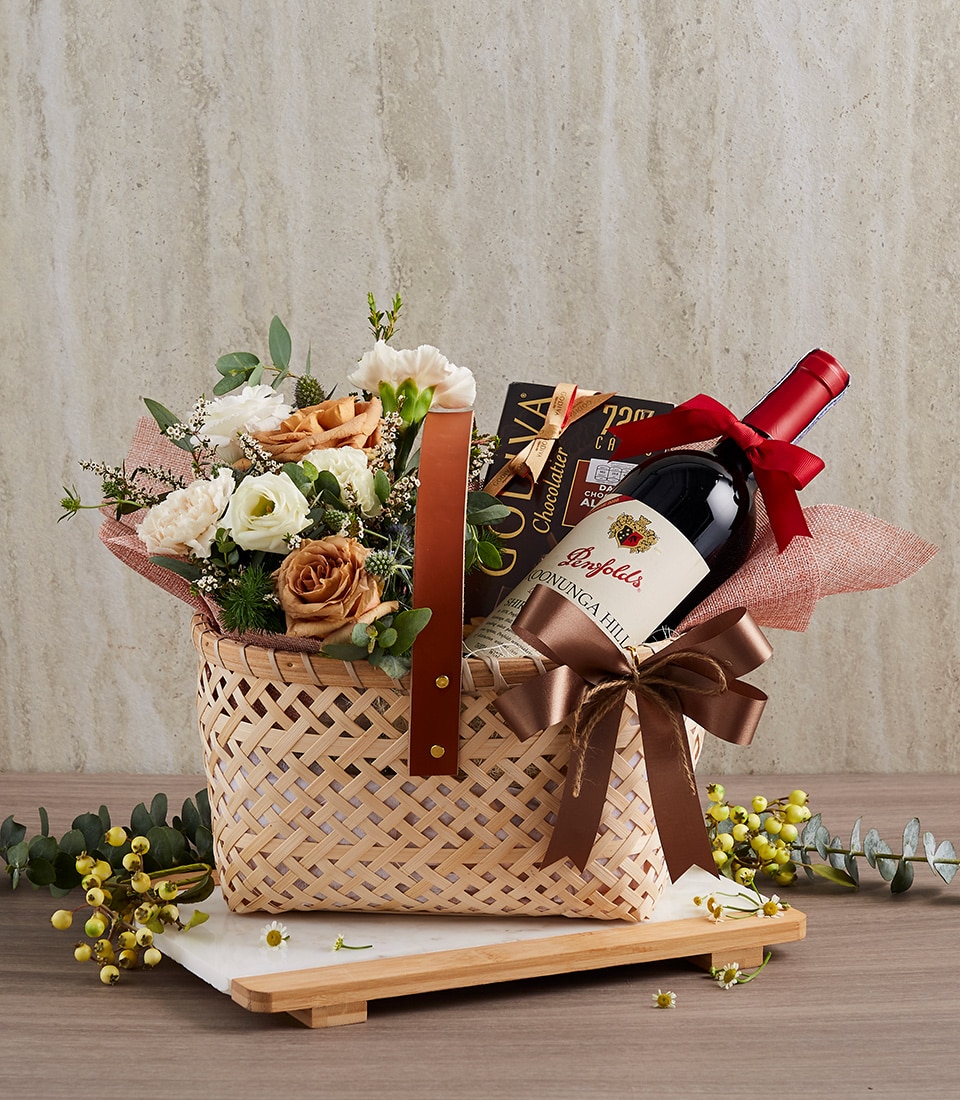 A luxurious woven basket adorned with the Basketeer logo, elegantly presented on a polished wooden surface. Inside, a bottle of exquisite red wine is wrapped in a rich brown ribbon, accompanied by a decadent bar of dark chocolate. An arrangement of vibrant fresh flowers, including lush roses and verdant greenery, adds an inviting touch. Delicate yellow buds are artfully scattered around the scene, enhancing the overall opulence and charm.