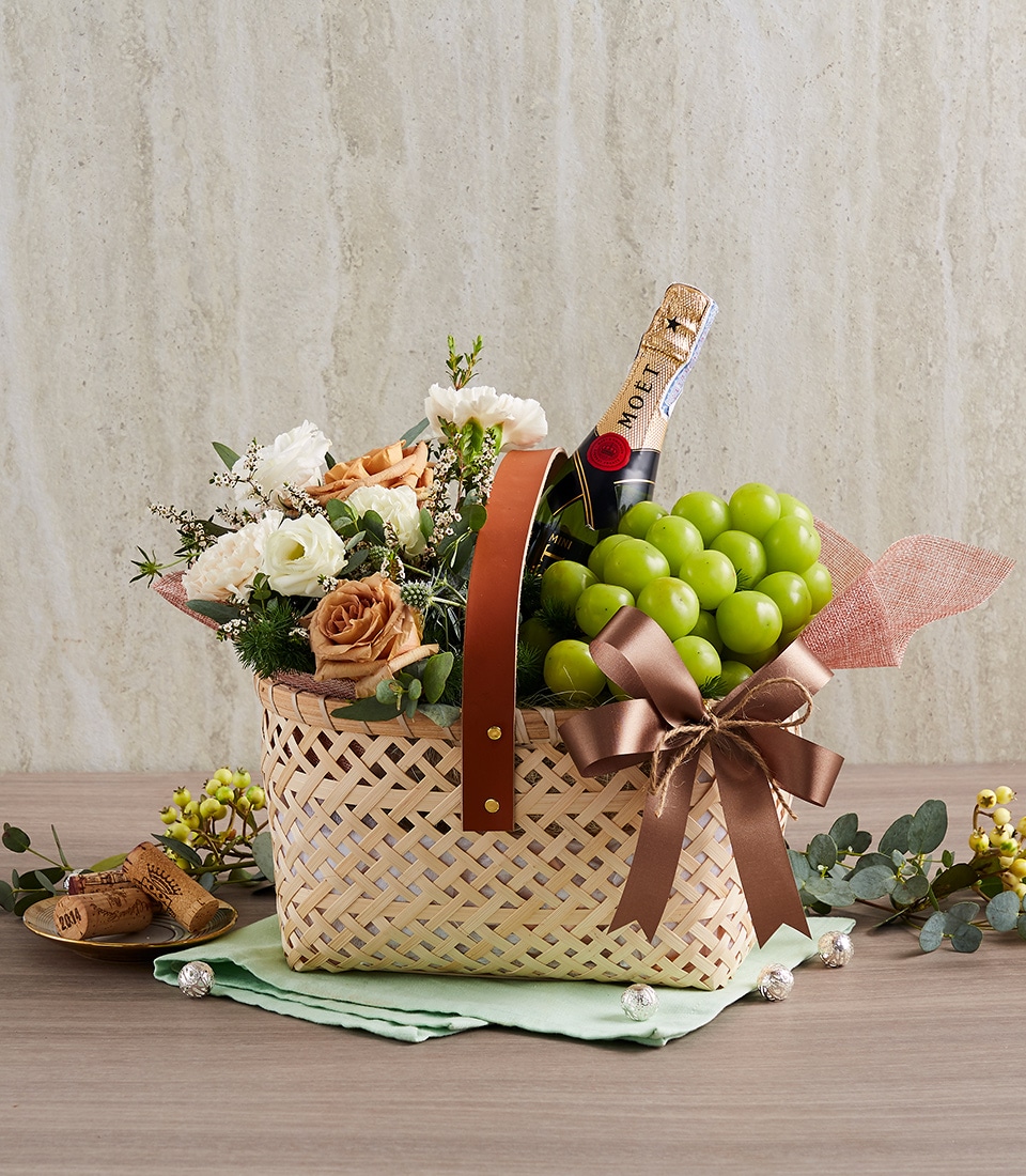 A lavish woven basket from Basketeer graces a polished wooden surface, brimming with opulence. Inside, a bottle of premium champagne nestles beside succulent green grapes, exquisite white and peach flowers, and lush greenery. The basket is adorned with an elegant leather handle and features a sophisticated brown ribbon bow bearing the Basketeer logo. Scattered across the table are delicate white blossoms and additional grapes, enhancing the luxurious scene with their natural charm.