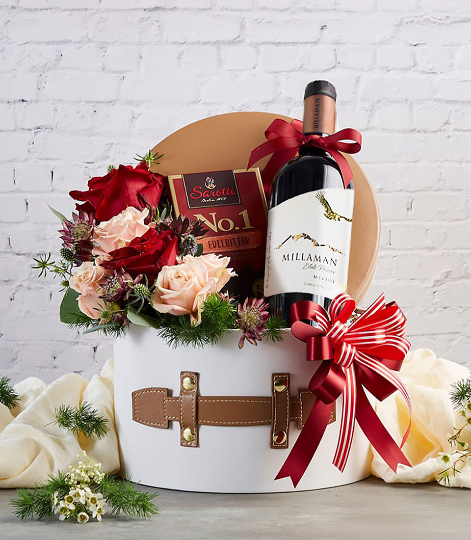 A premium gift box featuring a bottle of fine red wine, gourmet dark chocolate, and a stunning arrangement of red and blush roses, elegantly wrapped in a white hatbox with faux leather details and a red ribbon bow—perfect for romantic occasions, corporate gifts, and special celebrations.