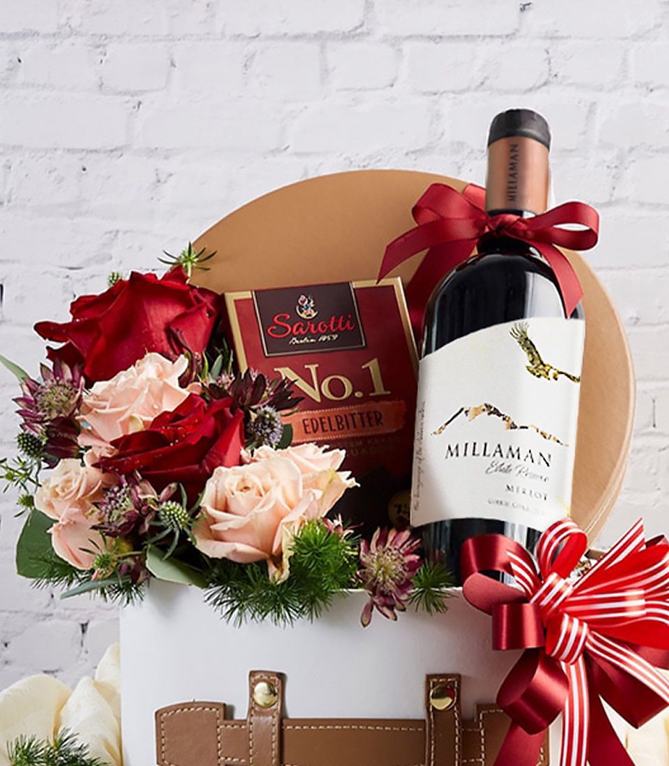 Close-up of a premium gift box featuring a bottle of fine red wine, gourmet dark chocolate, and a stunning arrangement of red and blush roses, elegantly wrapped in a white hatbox with faux leather details and a red ribbon bow—perfect for romantic occasions, corporate gifts, and special celebrations.