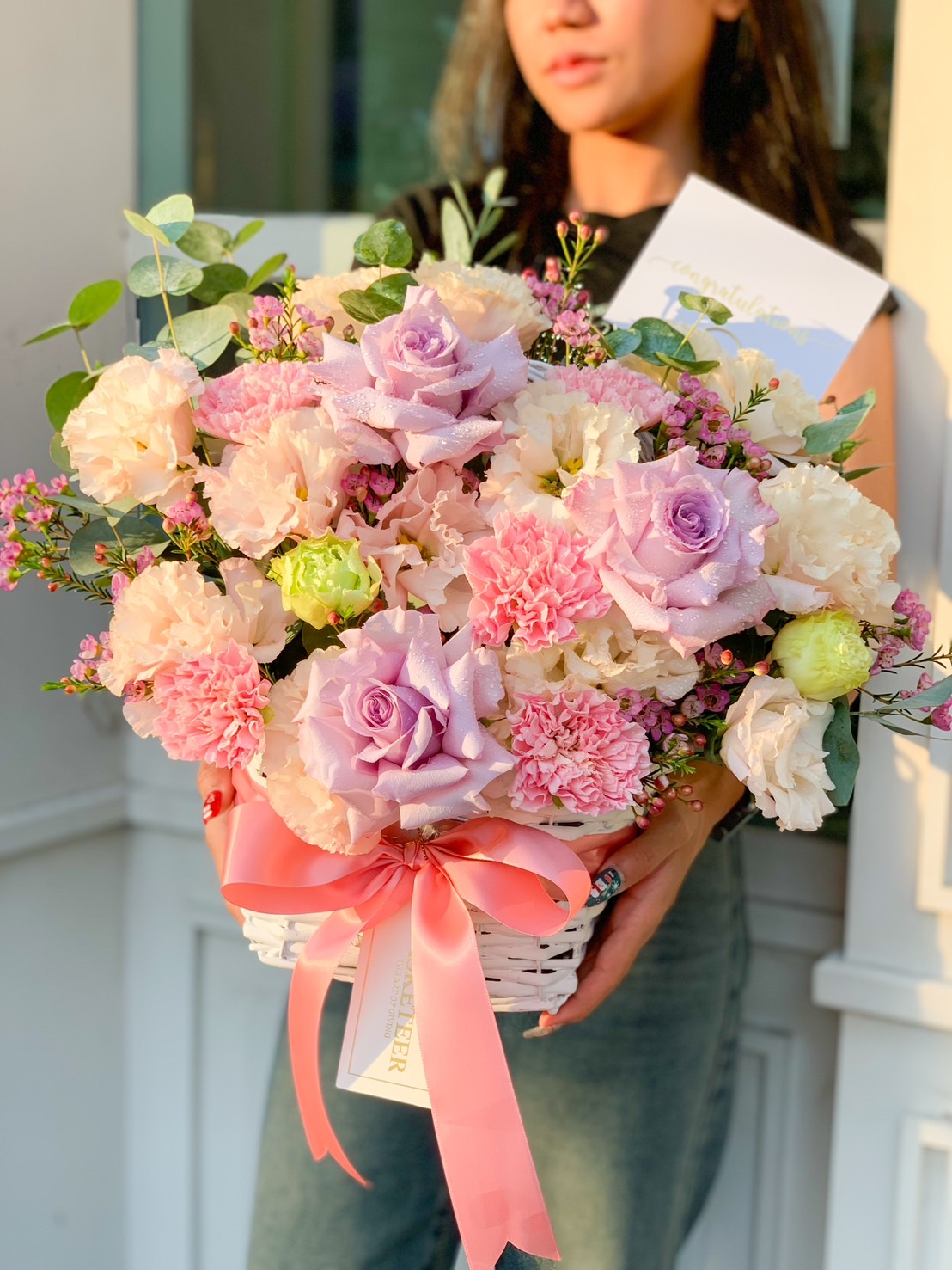 An elegant white wicker basket from Basketeer showcases a luxurious assortment of pastel blooms, featuring delicate pink, purple, and white roses alongside charming pink carnations and lush greenery. Adorned with an exquisite pink ribbon bow bearing the Basketeer logo, this arrangement rests on a pristine white marble surface against a sophisticated white paneled wall, creating an inviting and opulent display.
