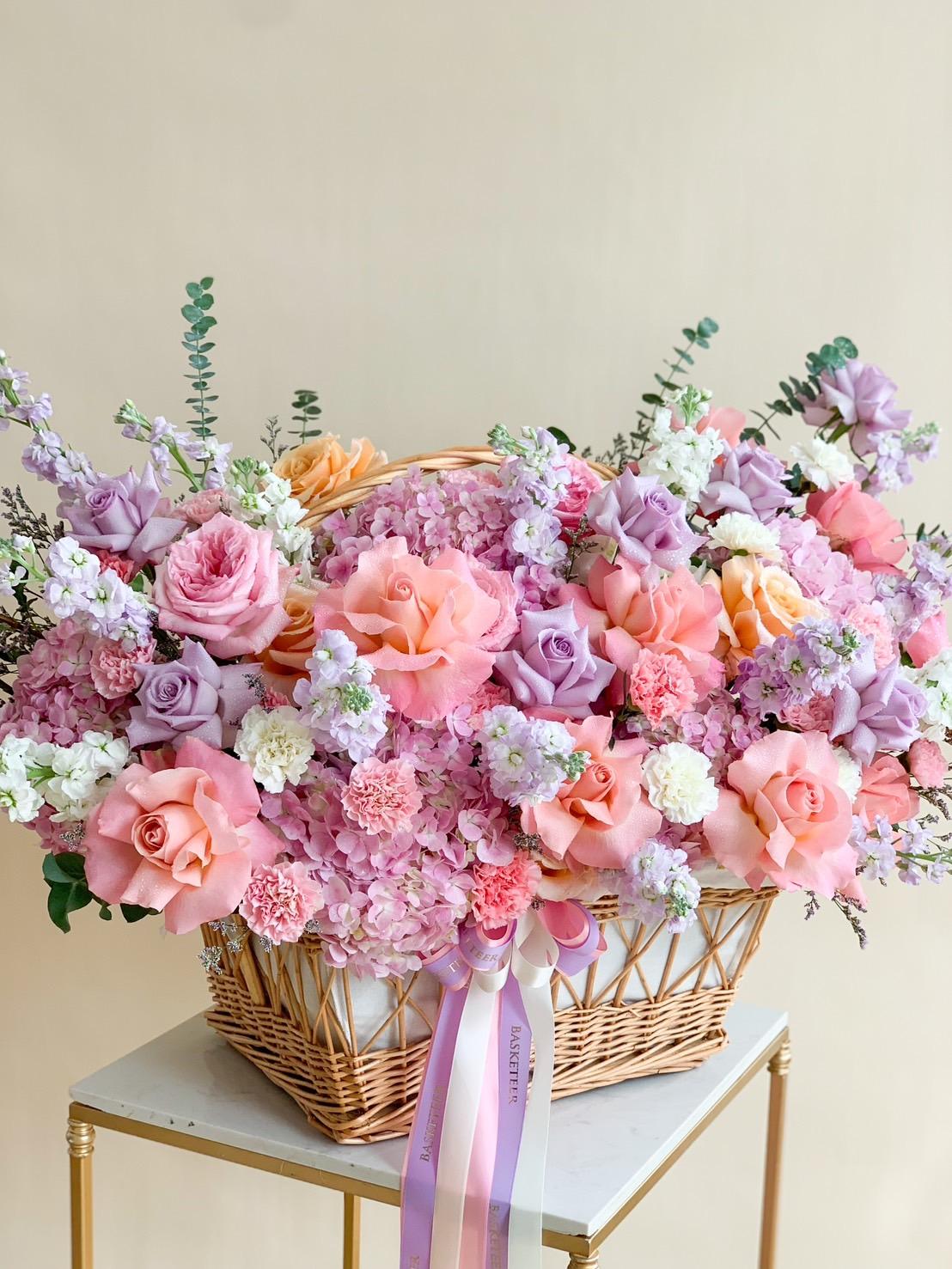 A wicker basket overflowing with a vibrant arrangement of pink, peach, and lavender roses, white and pink hydrangeas, and accented with sprigs of greenery. A pink ribbon with text is tied around the basket’s handle, all placed on a small, elegant table.
