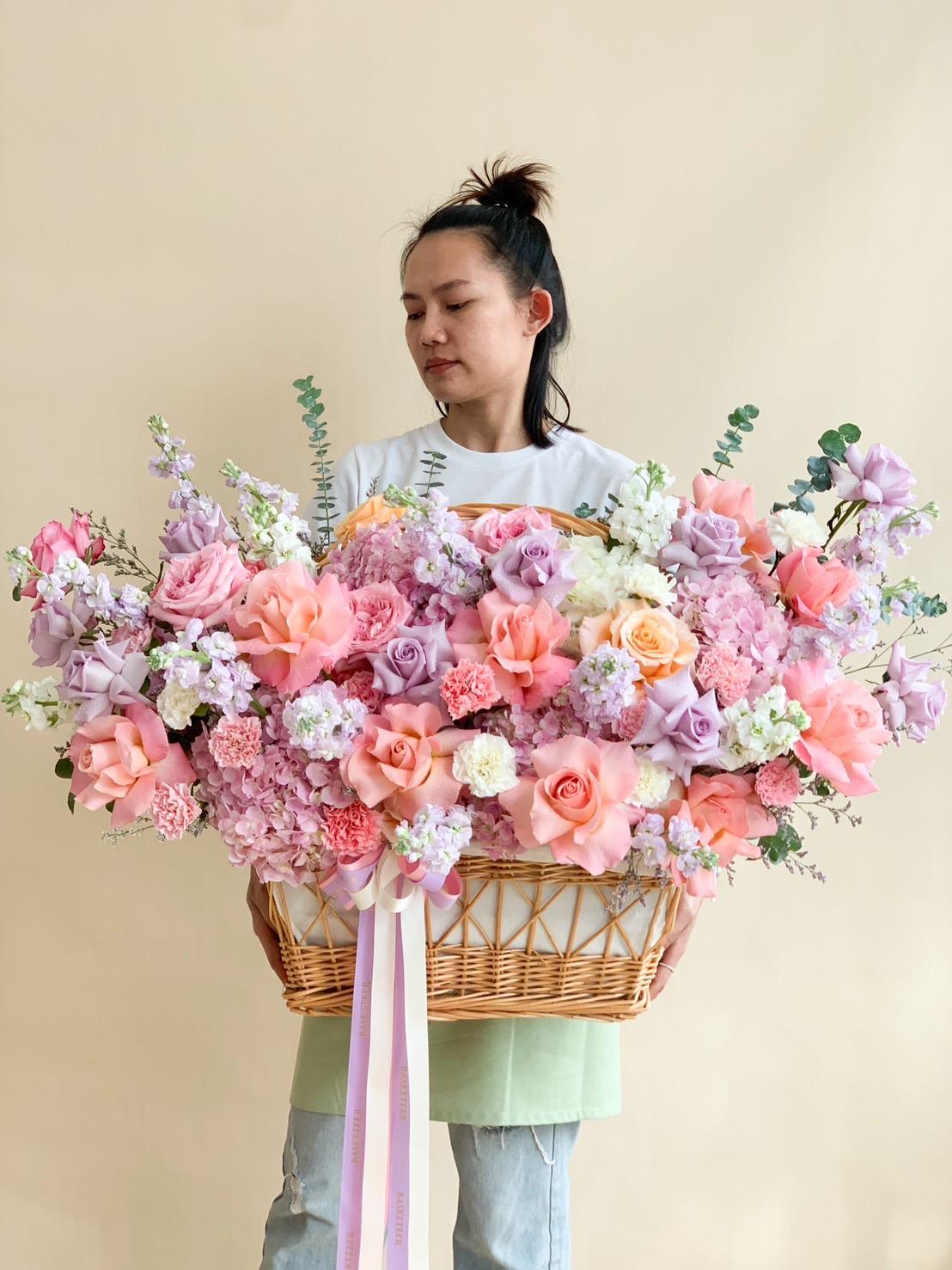 A person with tied-up hair holds a large, rectangular wicker basket overflowing with various pink, peach, and lavender flowers, adorned with greenery and a light purple ribbon cascading down the front. The background is a plain beige wall.