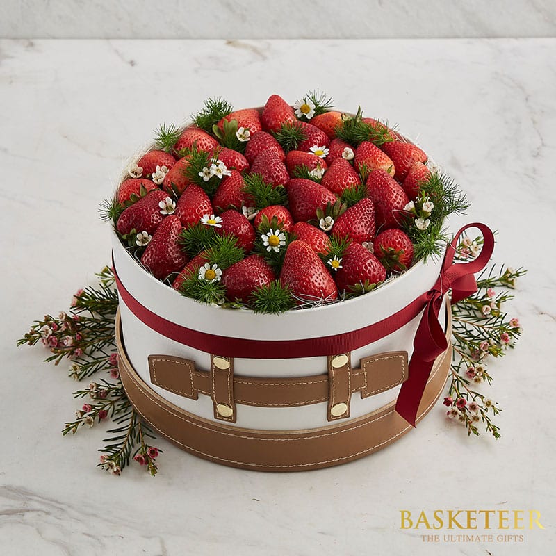 A luxurious round basket brimming with plump, fresh strawberries and delicate white flowers, elegantly accented with aromatic pine sprigs. The basket is tastefully wrapped in a pristine white cover with sophisticated brown leather-like straps and adorned with a vibrant red ribbon tied in an exquisite bow. The base of this delightful Fresh Strawberry Gift by Basketeer is further enhanced with lush greenery and charming floral accents, making it the perfect indulgence for any discerning recipient.