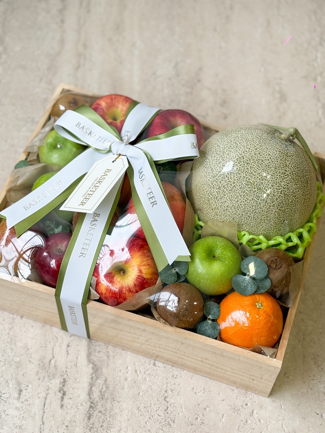 Experience Ultimate Fruit Box Bliss with this wooden gift box, brimming with apples, a melon, green apples, kiwi, tangerine, and grapes—all beautifully adorned with light green and white BASKETEER ribbon bows.