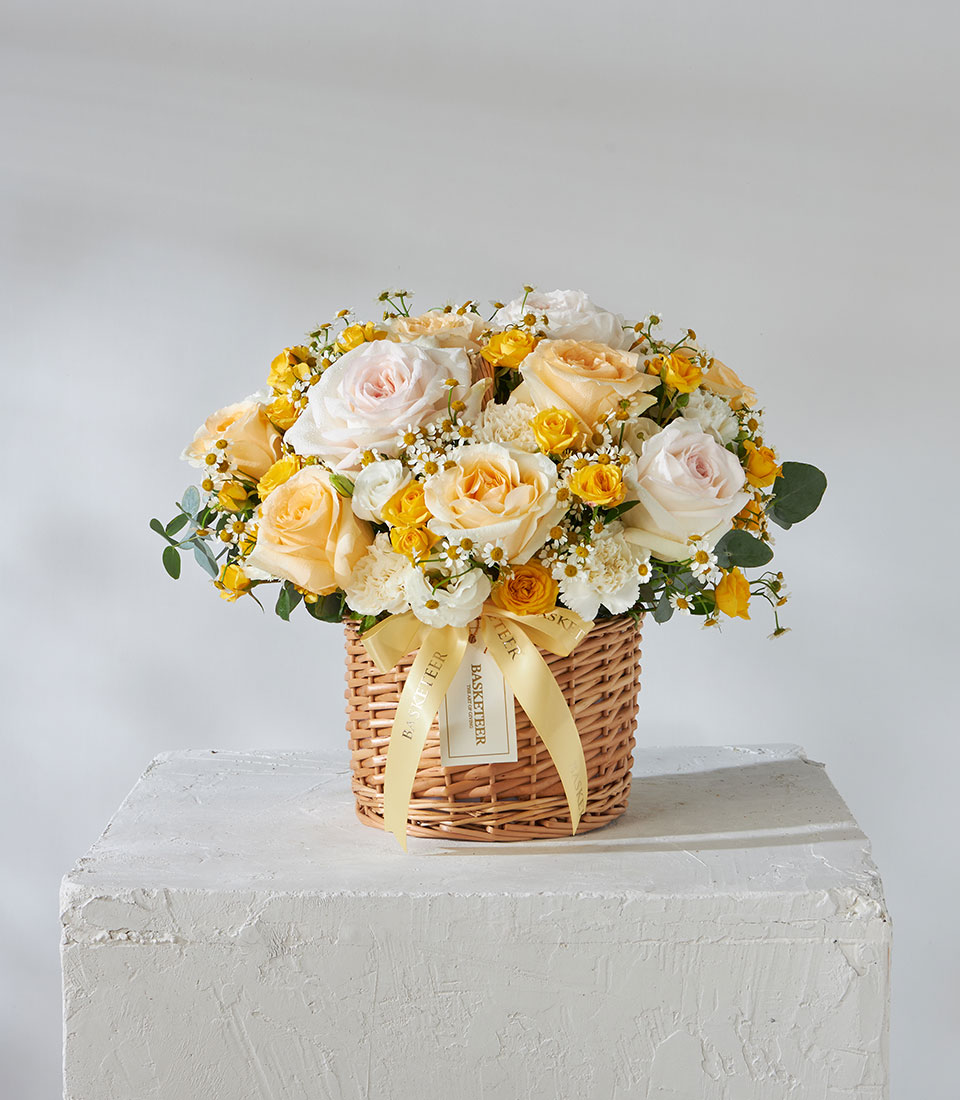 ตะกร้า Sunshine Bloom ประกอบไปด้วยดอกกุหลาบสีขาว เหลืองอ่อน และเหลือง พร้อมด้วยดอกไม้สีขาวเล็กๆ และใบไม้ในตะกร้าหวายบนฐานสีขาวที่มีพื้นผิวสัมผัส ตกแต่งด้วยริบบิ้นสีเหลืองและข้อความ