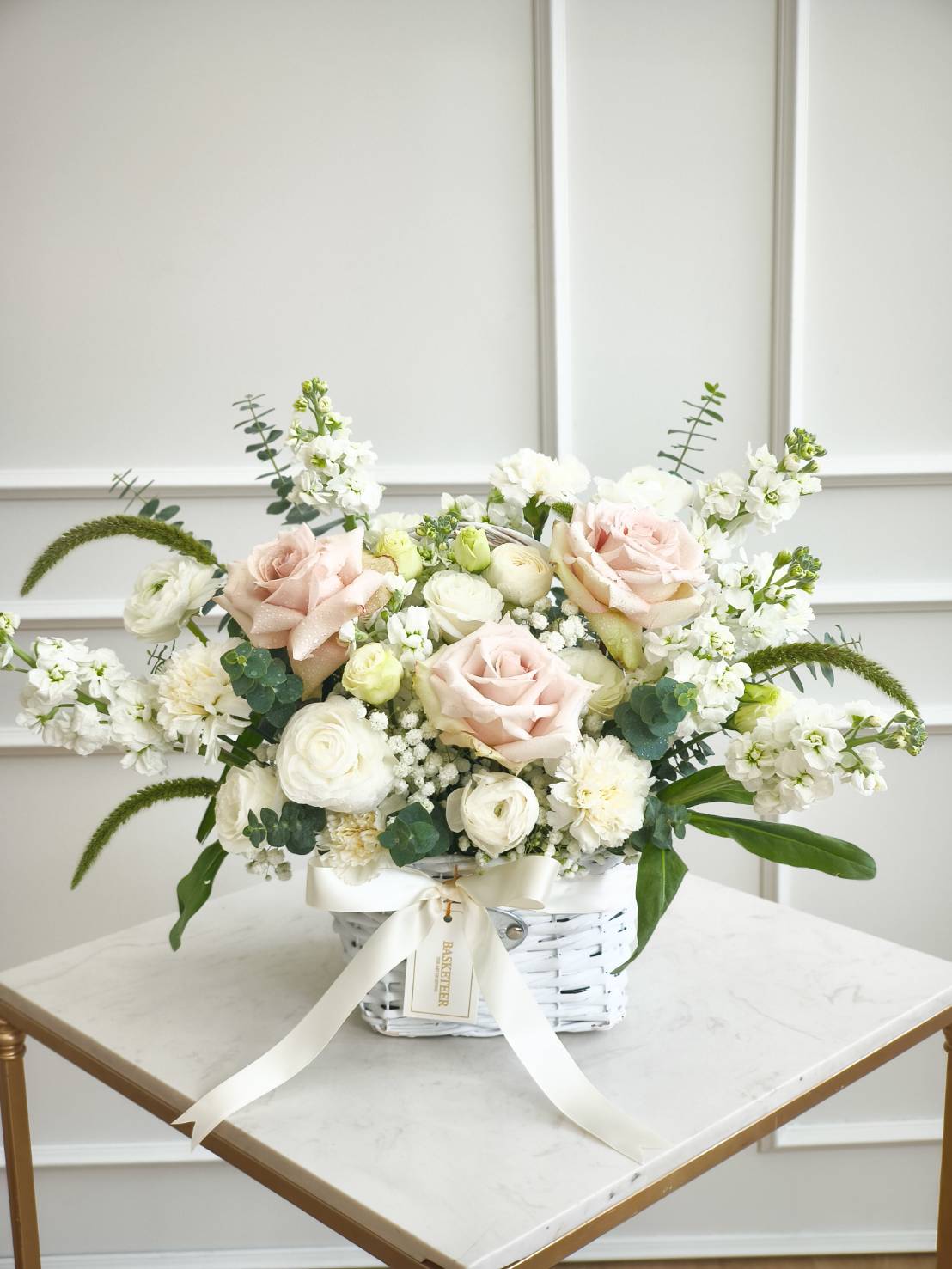 Elegant flower basket with ivory and blush roses, white ranunculus, carnations, and stock flowers, arranged with eucalyptus and white ribbon