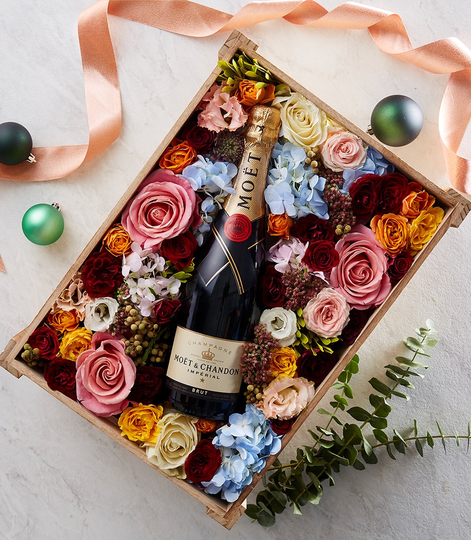 In an exquisitely crafted wooden box, a bottle of Moët & Chandon champagne takes center stage amidst a lavish display of vibrant roses and hydrangeas. Decorative green baubles add a touch of festive glamour, while a luxurious peach-colored ribbon adorned with the Basketeer logo enhances the opulent presentation. Set against a light, inviting background, this decadent arrangement from Basketeer exudes elegance and sophistication.