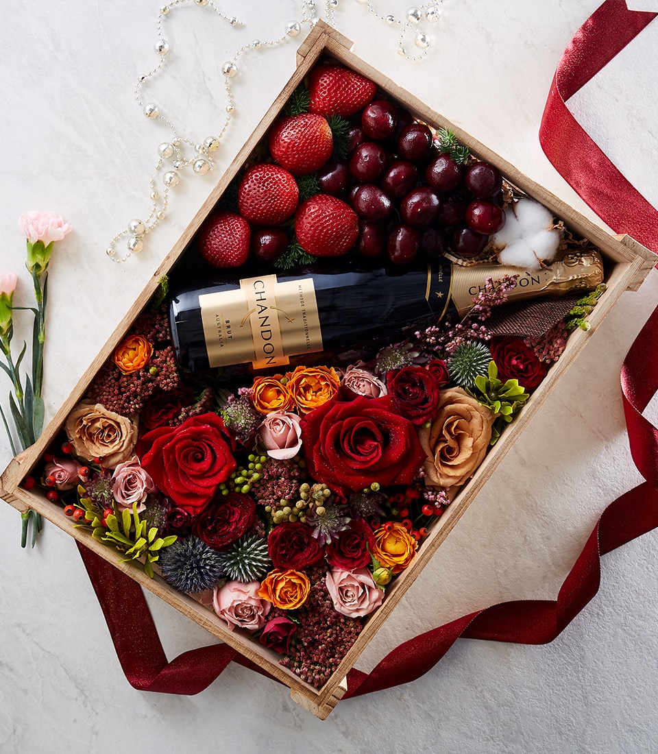 A lavish wooden Basketeer box showcases a bottle of Chandon champagne, nestled amidst an exquisite arrangement of vibrant red, pink, and yellow roses. Accented by luscious berries, verdant foliage, ripe strawberries, and sumptuous grapes, the display radiates luxury. A rich red ribbon adorned with the elegant Basketeer logo gracefully drapes around the box's edges, adding an extra layer of opulence to this inviting scene.