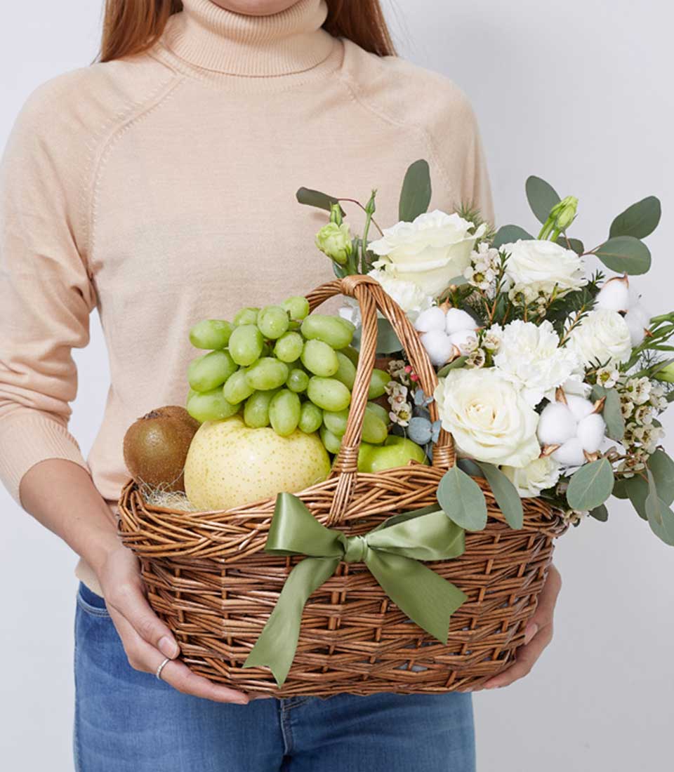 A person holds a wicker basket filled with green grapes, a round yellow fruit, a kiwi, and a variety of white flowers and green foliage. The Fruit & Flower Wonder Basket is adorned with a green ribbon tied in a bow. The person is wearing a light beige turtleneck sweater.