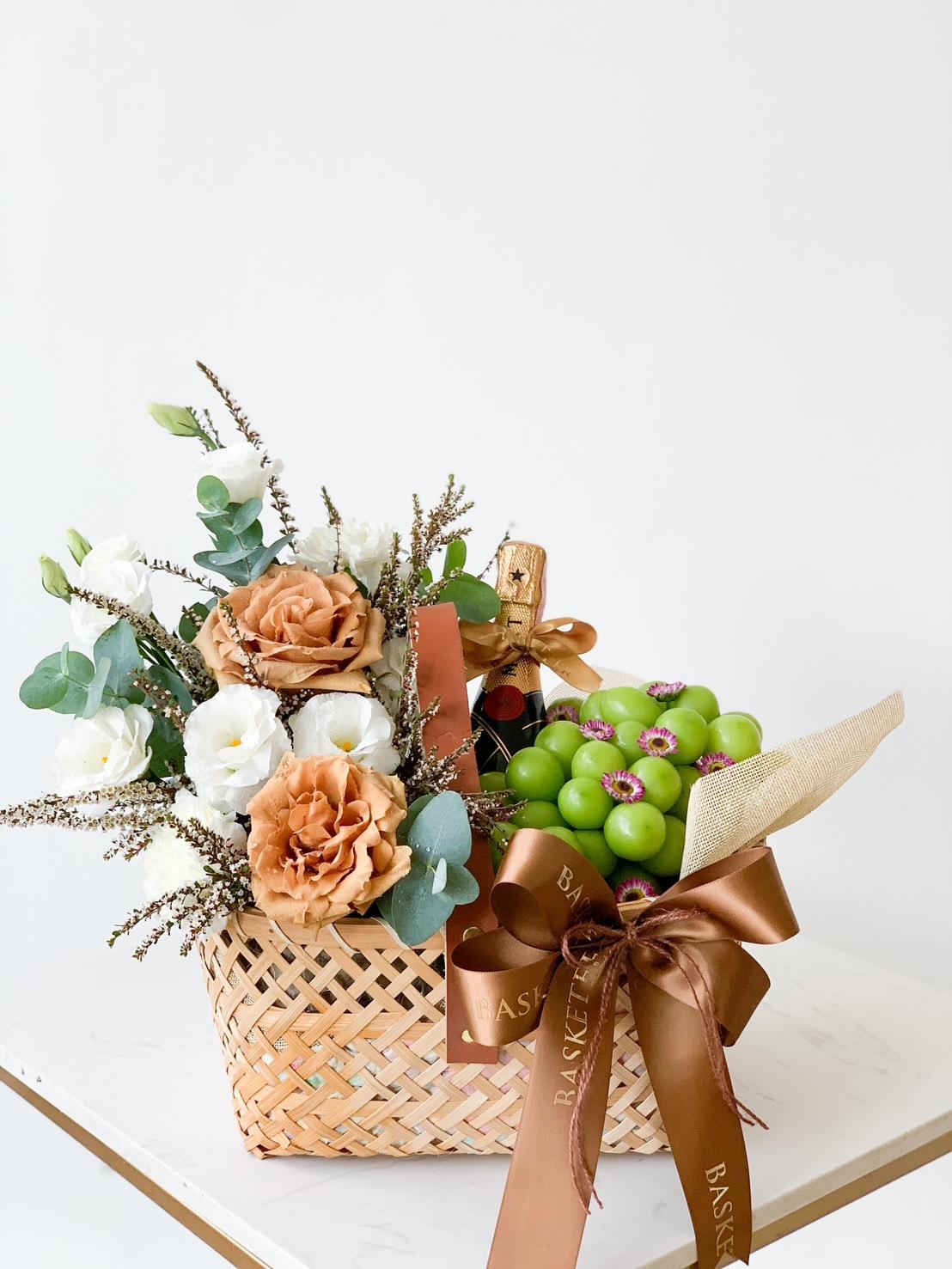 A wicker basket on a white surface contains a bouquet of orange and white flowers, greenery, a cluster of green grapes, and a wrapped bottle. The basket is adorned with brown ribbon bows and has a tag reading "BASKET" attached to one of the ribbons.