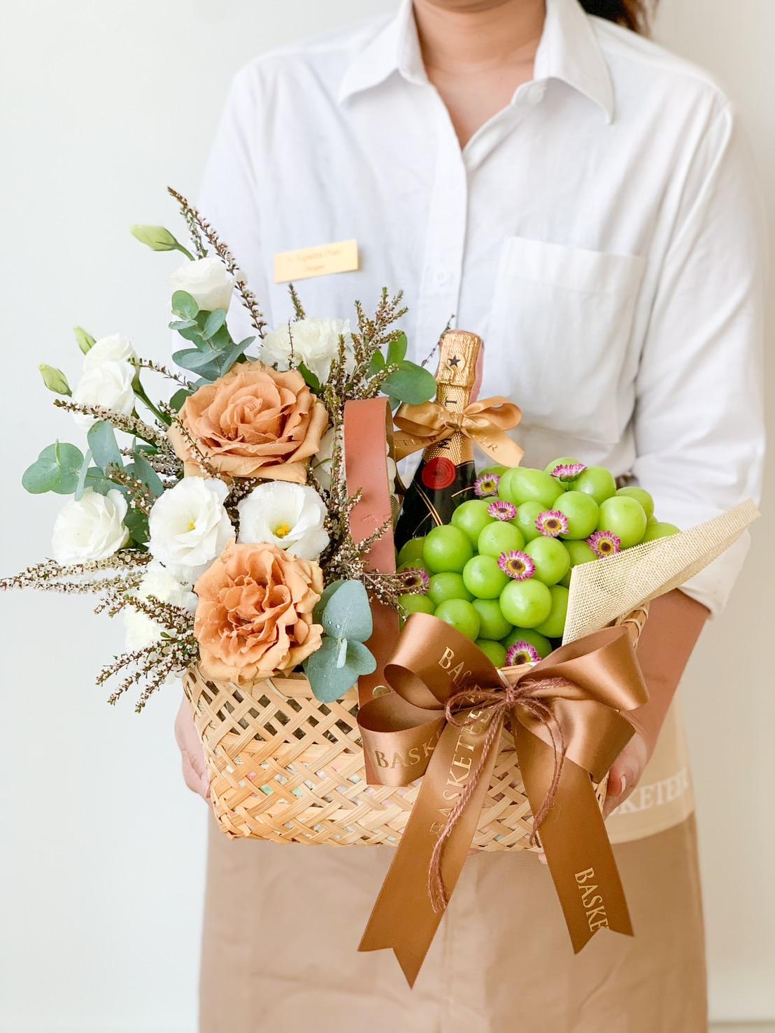 A person in a white shirt and beige apron holds a wicker basket containing orange and white flowers, a bottle of champagne, and a bunch of green grapes. The basket is adorned with brown ribbons that have "Basket" printed on them.