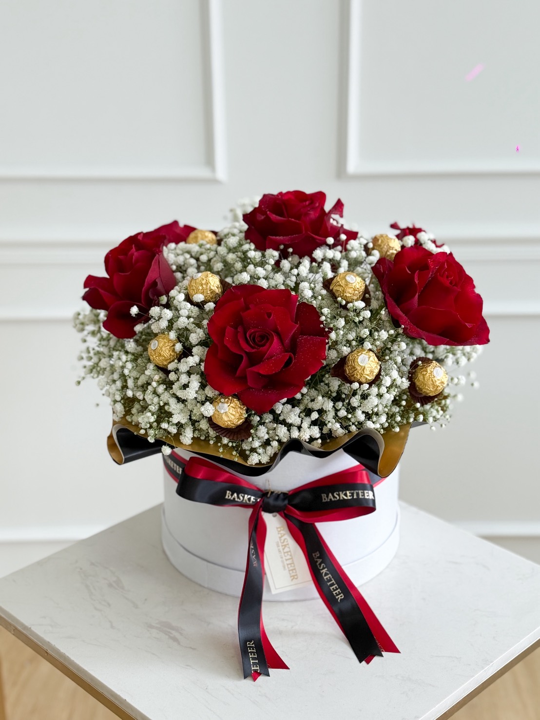 A Luxury Roses & Ferrero Rocher Gift Box filled with red roses, baby’s breath, and gold-wrapped chocolates sits on a white marble table, decorated with black and red ribbons against a white panelled wall.