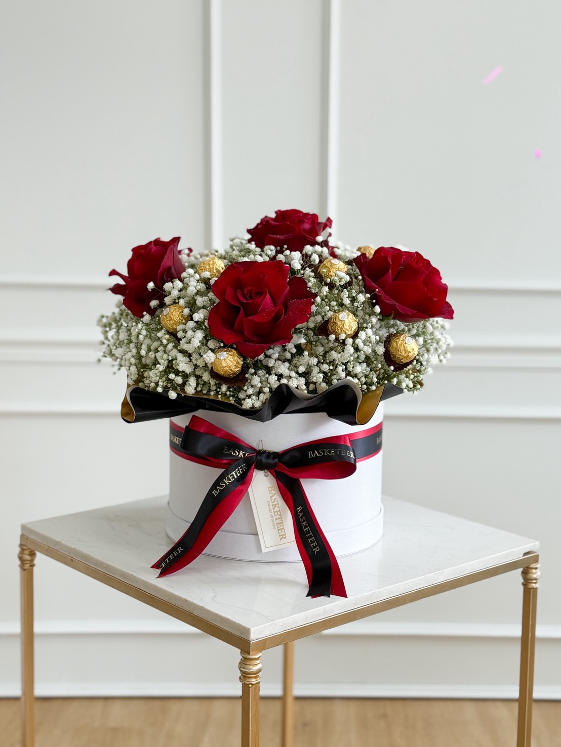 A round white Luxury Roses & Ferrero Rocher Gift Box with a black and red ribbon holds red roses, white baby's breath, and gold-wrapped chocolates, set on a small white table with gold legs against a panelled wall.