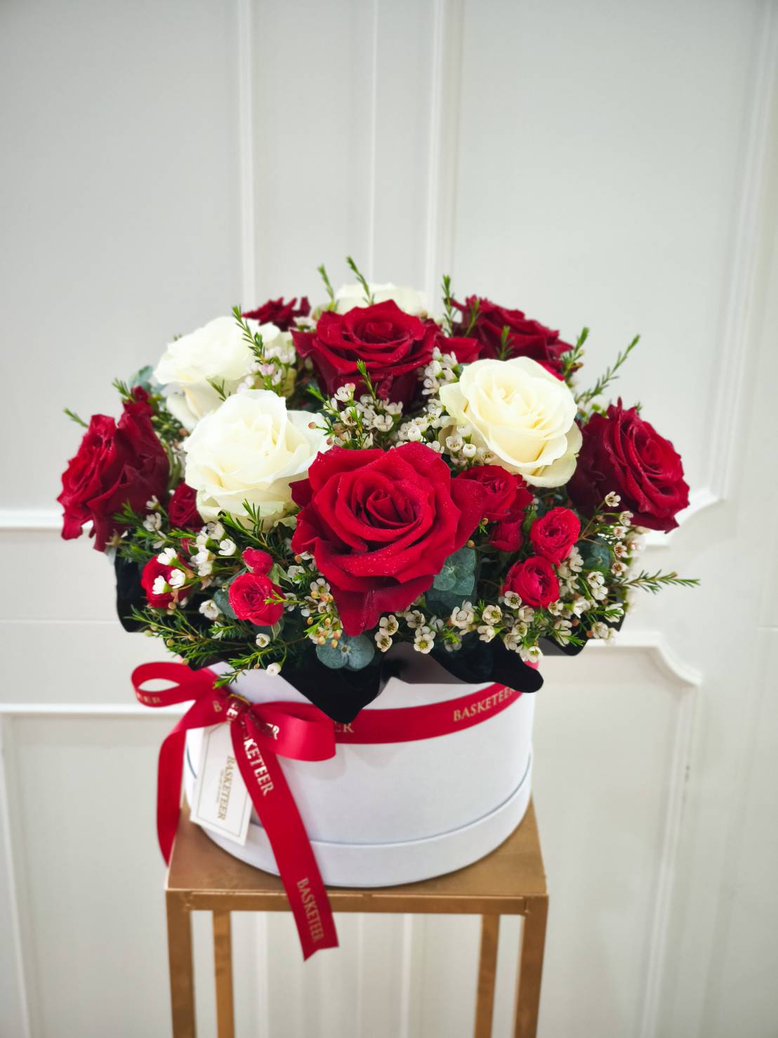 Round white box arrangement of red roses, white roses, and waxflowers, decorated with a red ribbon.