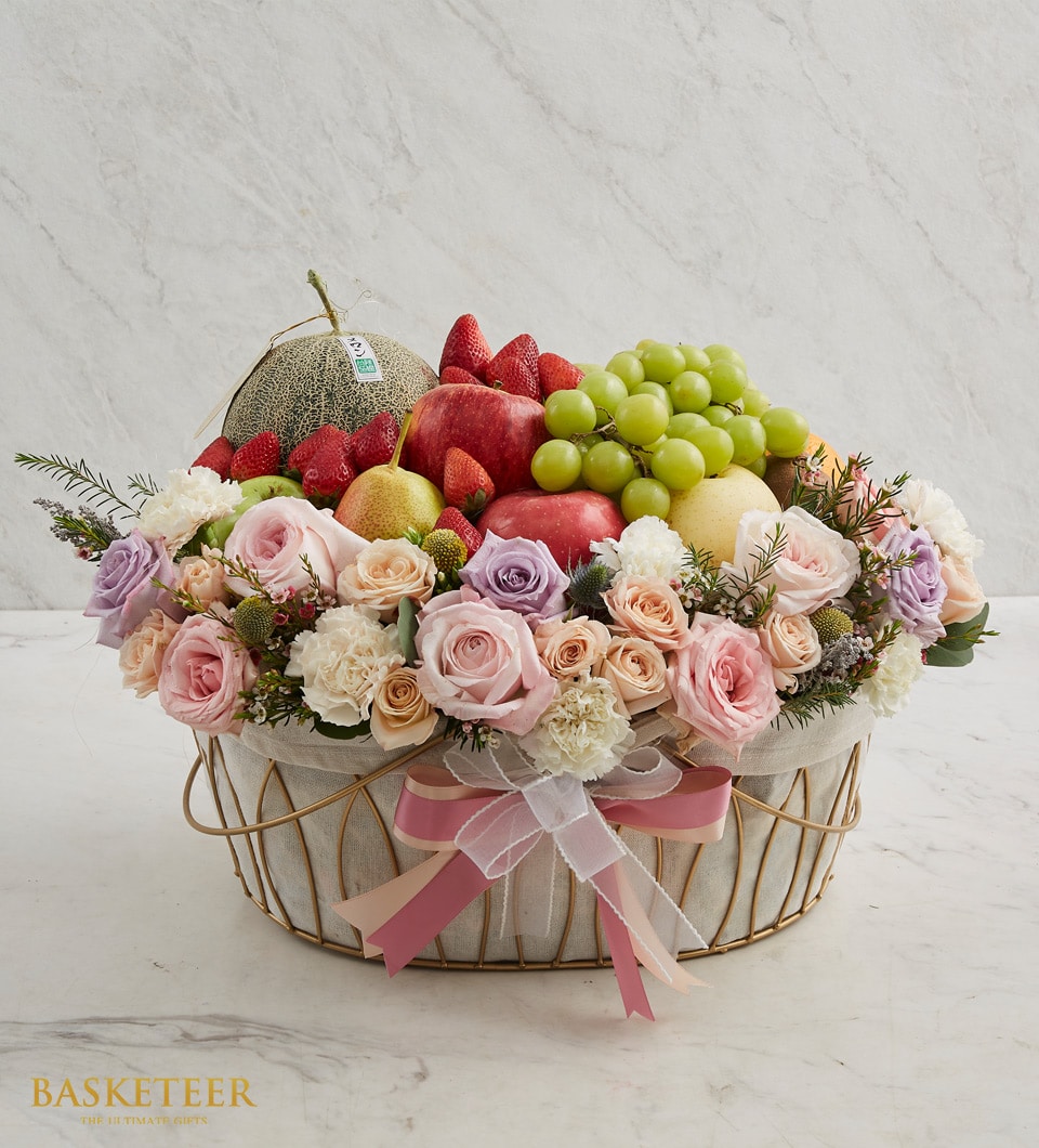 An opulent Harvest Fruit Blossom Basket brimming with an exquisite assortment of fresh fruits, including a succulent melon, luscious strawberries, crisp apples, and vibrant green grapes. This bountiful selection is beautifully complemented by a stunning arrangement of colorful roses and carnations. The basket is elegantly adorned with a luxurious ribbon and proudly displays the 