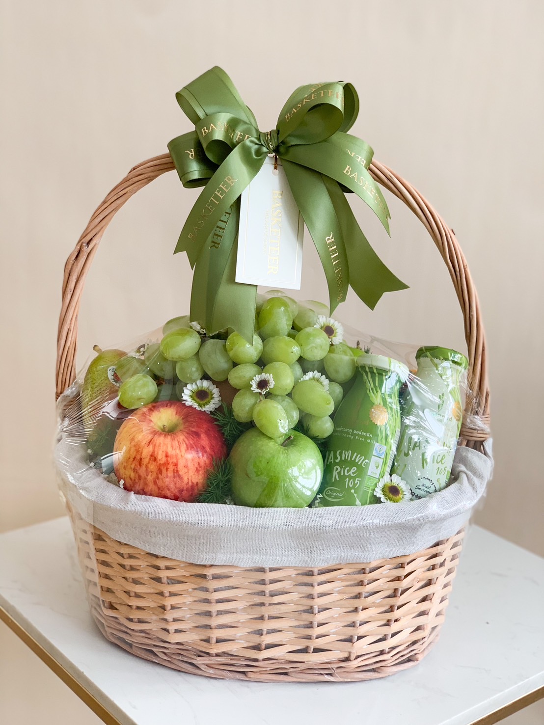 A white rattan gift basket with fabric lining, filled with fresh fruits such as green grapes and red apples, healthy drinks, and decorated with green ribbon bows.