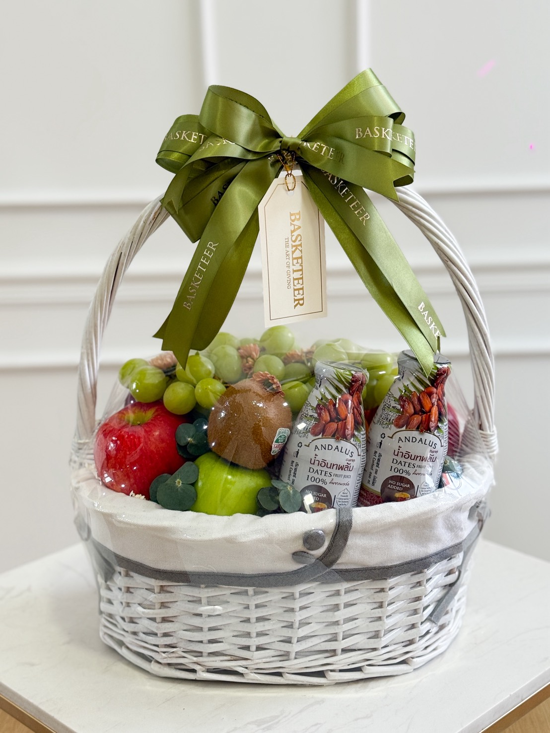 A white wicker Wonder Fruit Gift Hamper filled with assorted fresh fruit and two bottles of juice, wrapped in clear plastic and topped with a green ribbon and a 