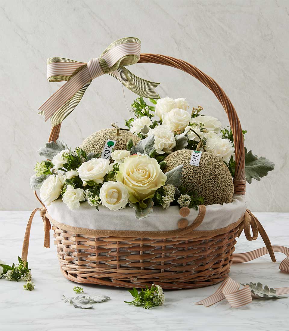 Natural brown rattan gift basket with fabric lining inside. Filled with fresh fruits and green melons With fresh white and green roses Decorated with green and light brown ribbon bows.