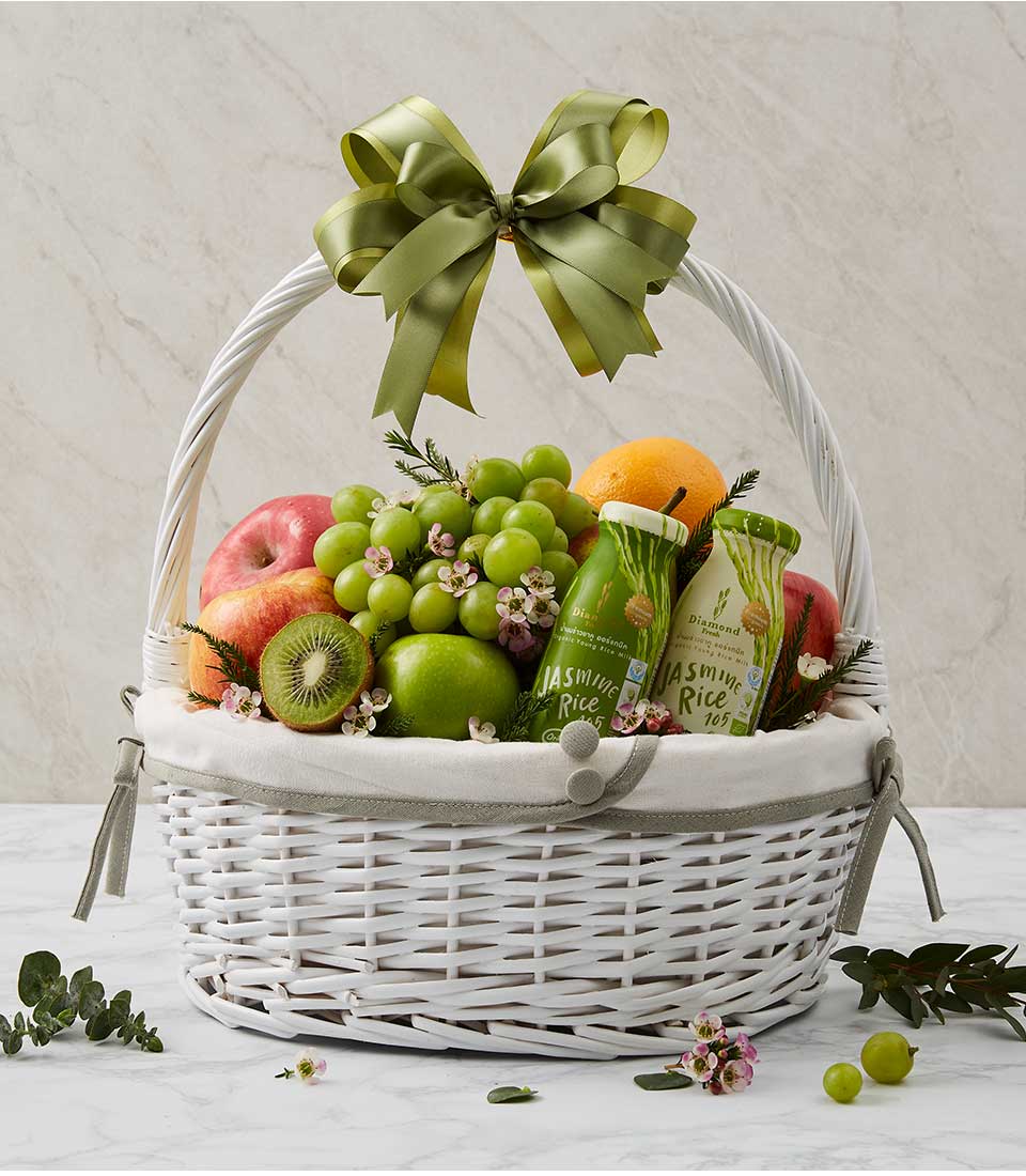 A white rattan gift basket with fabric lining, filled with fresh fruits such as green grapes and red apples, healthy drinks, and decorated with green ribbon bows.