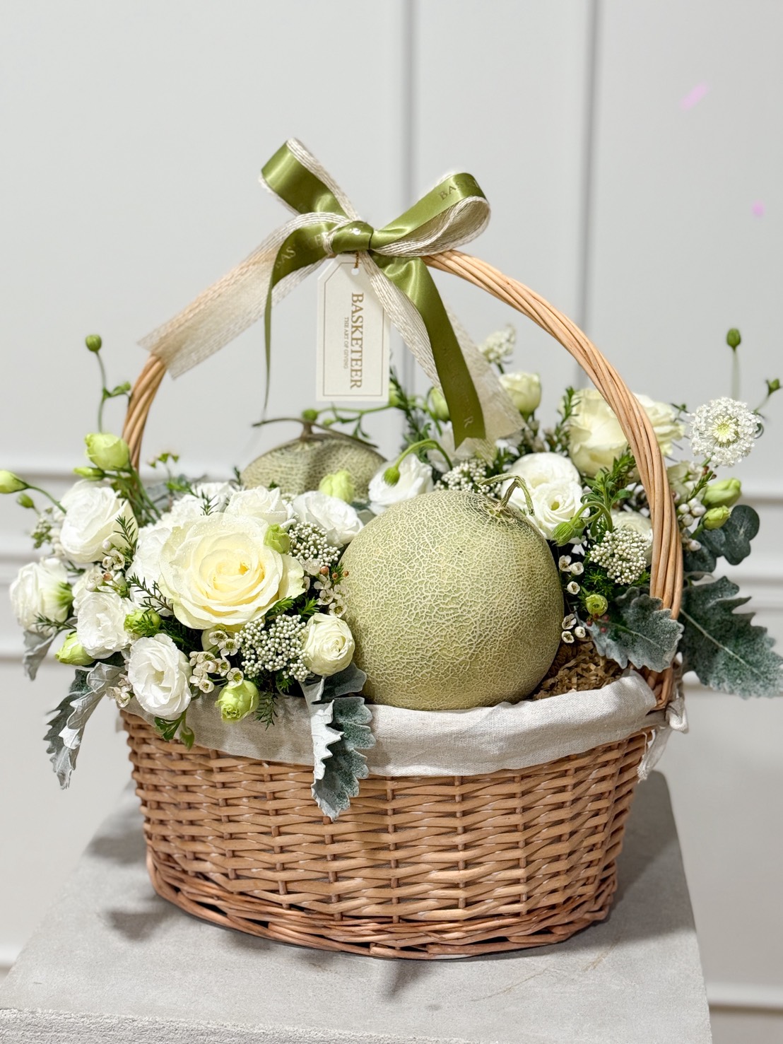 A Freshy Melon Gift Hamper featuring a wicker basket filled with white roses, greenery, and two cantaloupe melons, adorned with a green ribbon and tag, set against a plain light background.
