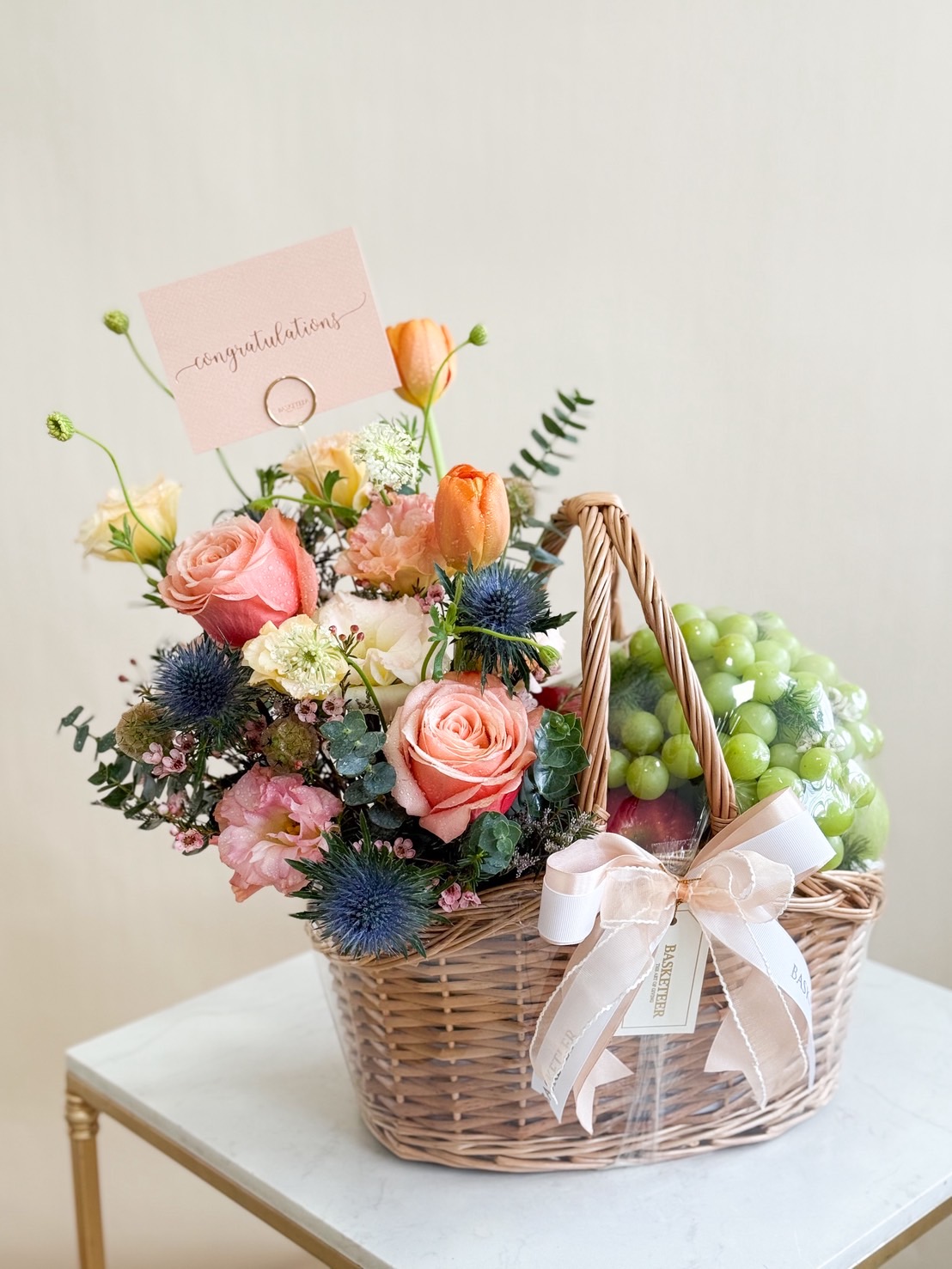 Elegant wicker basket filled with green grapes and apples, adorned with roses, tulips, carnations, eryngium, and delicate filler flowers.