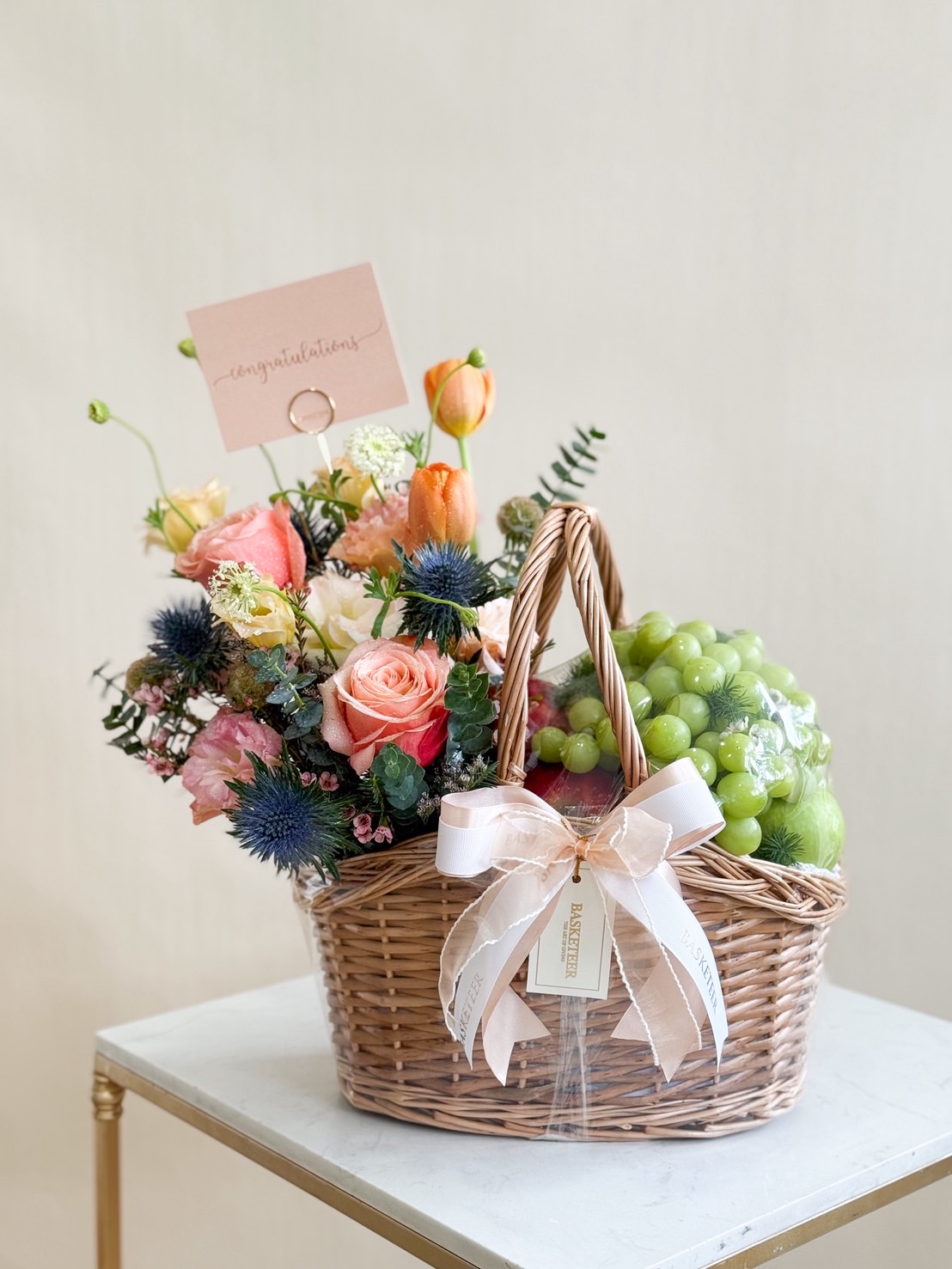 Natural wicker basket filled with green grapes and apples, decorated with roses, tulips, carnations, eryngium, and lush greenery.