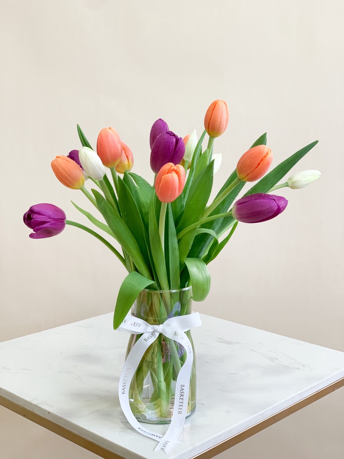 Colourful tulips in purple, orange, and white beautifully arranged in a clear glass vase with white ribbon.
