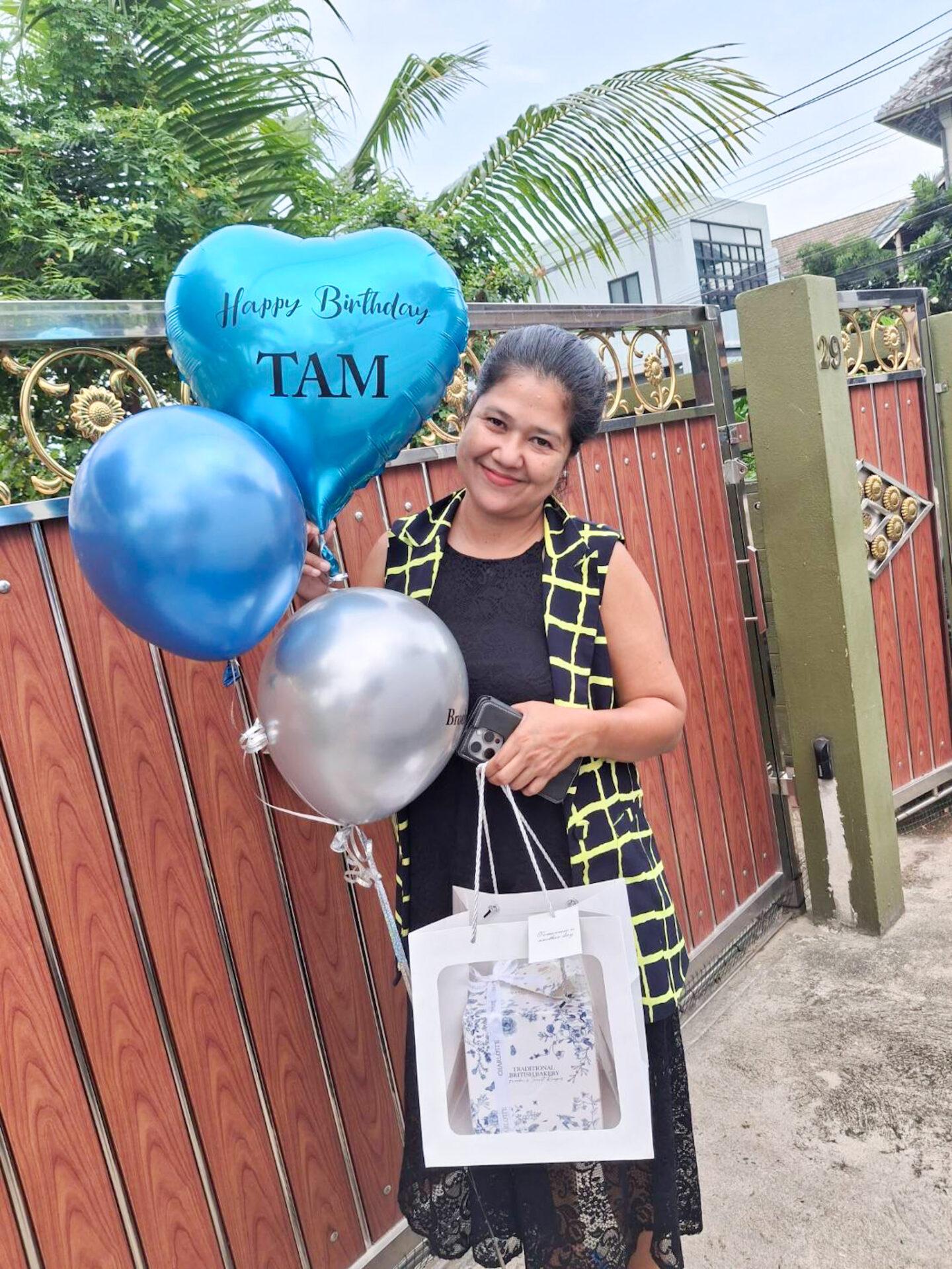 A woman wearing a black lace dress with a yellow grid-patterned vest is holding a Blue Heart Balloons Combo Gift Set and a gift bag. One blue balloon reads 