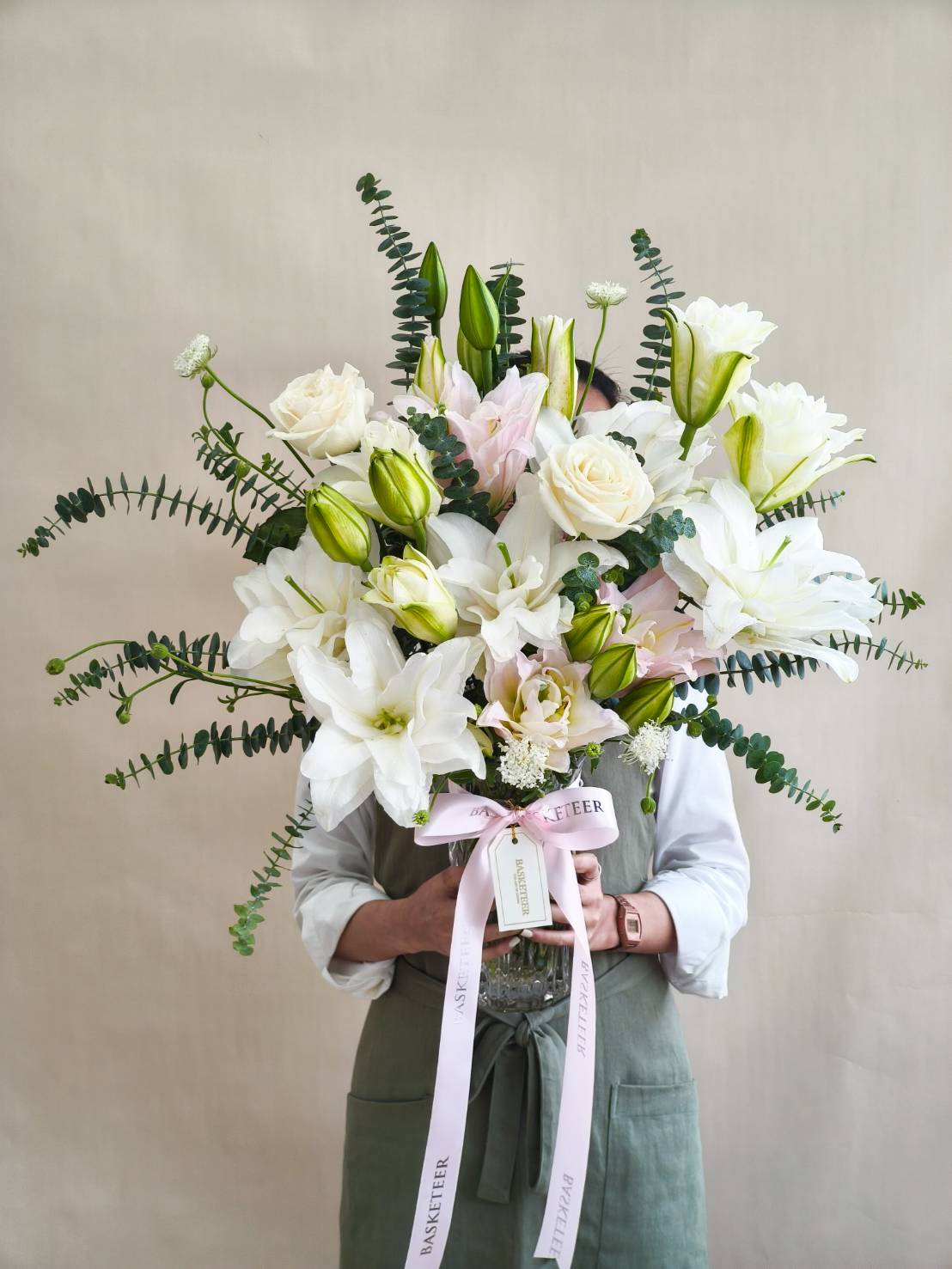 Luxurious glass vase with white lilies, ivory roses, pastel blooms, and eucalyptus greenery tied with a pink satin ribbon